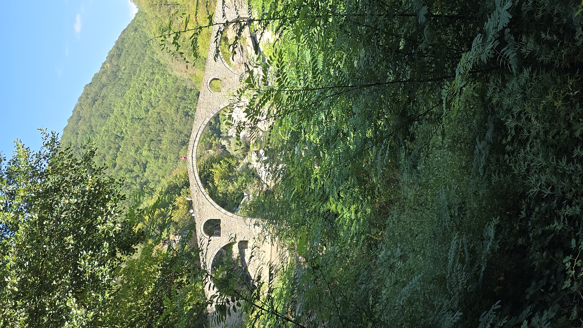 The image captures an ancient stone arched bridge situated in the village of Diadovtsi, Bulgaria. The bridge, constructed from weathered grey stone, features three prominent arches, with the central arch being the largest and most significant. Smaller, semicircular openings are visible within the structure, possibly for water flow or structural support. A few people, appearing as small figures from this distance, are visible walking on the bridge. The bridge spans across a rocky riverbed, with visible water flowing beneath. The surrounding environment is lush and verdant, dominated by dense green foliage and trees that frame the bridge. In the background, a steep, tree-covered mountainside rises towards a clear blue sky. The lighting suggests it is daytime, with sunlight illuminating the scene and casting shadows, contributing to the overall natural and historical ambiance.