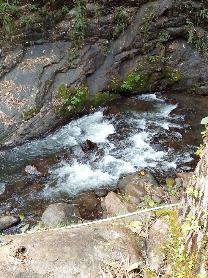 The image displays a natural stream flowing through a narrow, rocky channel within the city of Sukadana, Indonesia, captured on September 6, 2025, at 14:43. The water, appearing clear with visible rapids and turbulent white foam, navigates over numerous dark, rounded rocks embedded in the streambed. The left bank consists of a steep, dark grey rock face exhibiting distinct geological stratification, with sparse green moss and small plants growing in crevices and scattered dry leaves. The right bank in the foreground features a flatter, lighter grey/brown rock or concrete surface, also partially covered with moss and small green flora. A thin, horizontal white line, possibly a pipe or cable, runs along this lower right bank. Further right, a textured vertical surface, likely part of a tree or rock, is densely covered in moss and small ferns. No human subjects or their direct interactions are present. The scene is illuminated by natural light, indicating an outdoor, natural environment.