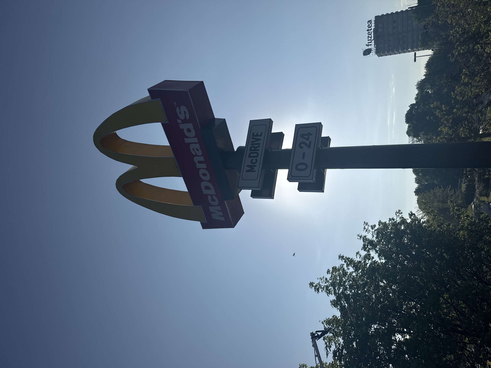 The image shows a McDonald's sign on a pole against a bright sky. The sign includes the golden arches, the McDonald's logo, and signs indicating "McDRIVE" and "0-24" (24 hours). In the background, a tall building with the "fuzetea" logo is visible. Trees are present on the right side of the frame. The location is Sofia, Bulgaria.