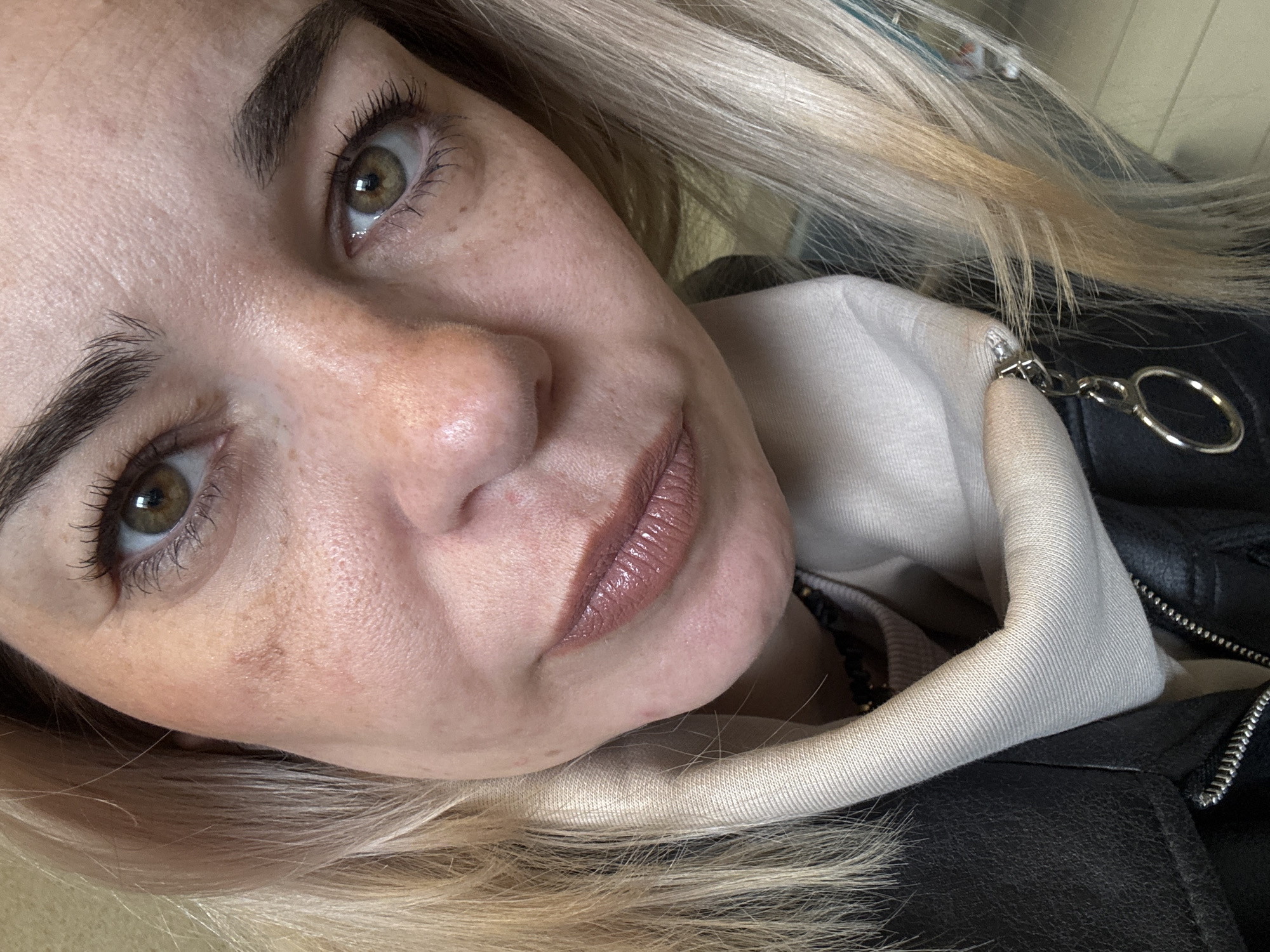 A close-up, portrait-style photograph depicts a fair-skinned woman with blonde hair. Her face is characterized by hazel eyes, dark eyebrows, freckles, and a lip color in a neutral, mauve shade. She is wearing a black leather jacket over a light-colored hooded sweatshirt. A small silver keychain is visible attached to the jacket. The setting and city cannot be determined from the provided image.