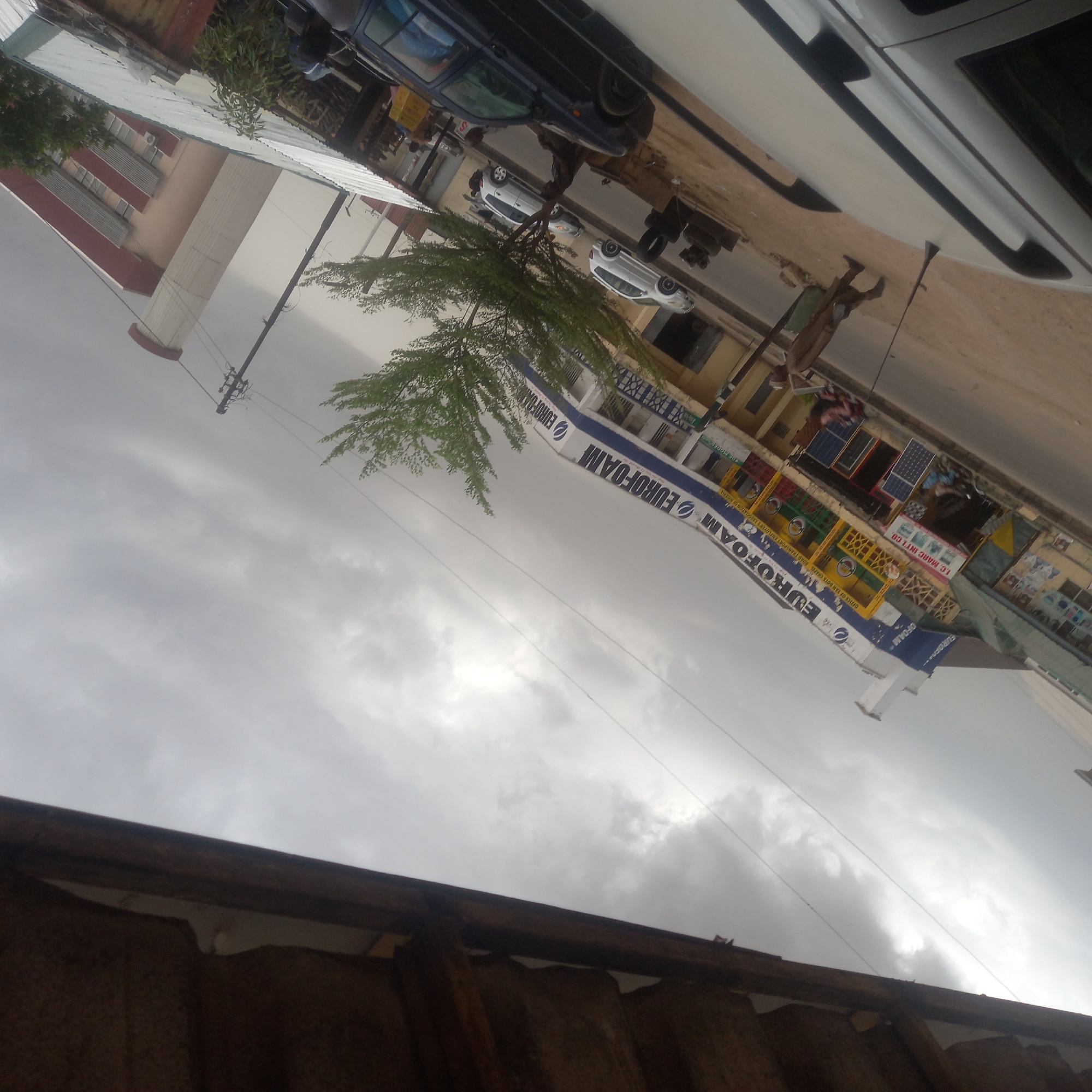 This media file captures an overcast daytime street scene in an urban or semi-urban area, likely Dadi Riba, Nigeria. The image is oriented vertically but depicts a horizontal street view.
The sky is heavily overcast with grey clouds, indicating a cloudy day. In the foreground, a dark, possibly corrugated metal roof is visible along the bottom edge.
The midground features a paved street with several vehicles. A dark blue SUV is partially visible in the upper right, while further down the street, two white cars are parked. Utility poles with power lines run alongside the road. Lush green trees are interspersed along the street, providing some shade.
Lining the street are various buildings, some multi-storied, housing shops and businesses. Visible signs and banners include a prominent white and blue banner with the text "EUROFOAM" repeated multiple times. Other businesses display merchandise, such as solar panels visible on display at one stall.
Several people are discernible on the street. One individual is seen standing near a white car, possibly attending to it or waiting. Others appear to be walking or standing near the shops, engaging in daily activities.
Additional visible text includes "1.C. MARC INT'L CO." and a partially visible sign that reads "OFFICE OF YAN DOTS CHAPEL ROAD TRANSPORT EMPLOYERS ASSOCIATION OF WO" (likely "OF NIGERIA"). Another fragment reads "DE MODEL". The overall impression is a bustling commercial area under a muted sky.