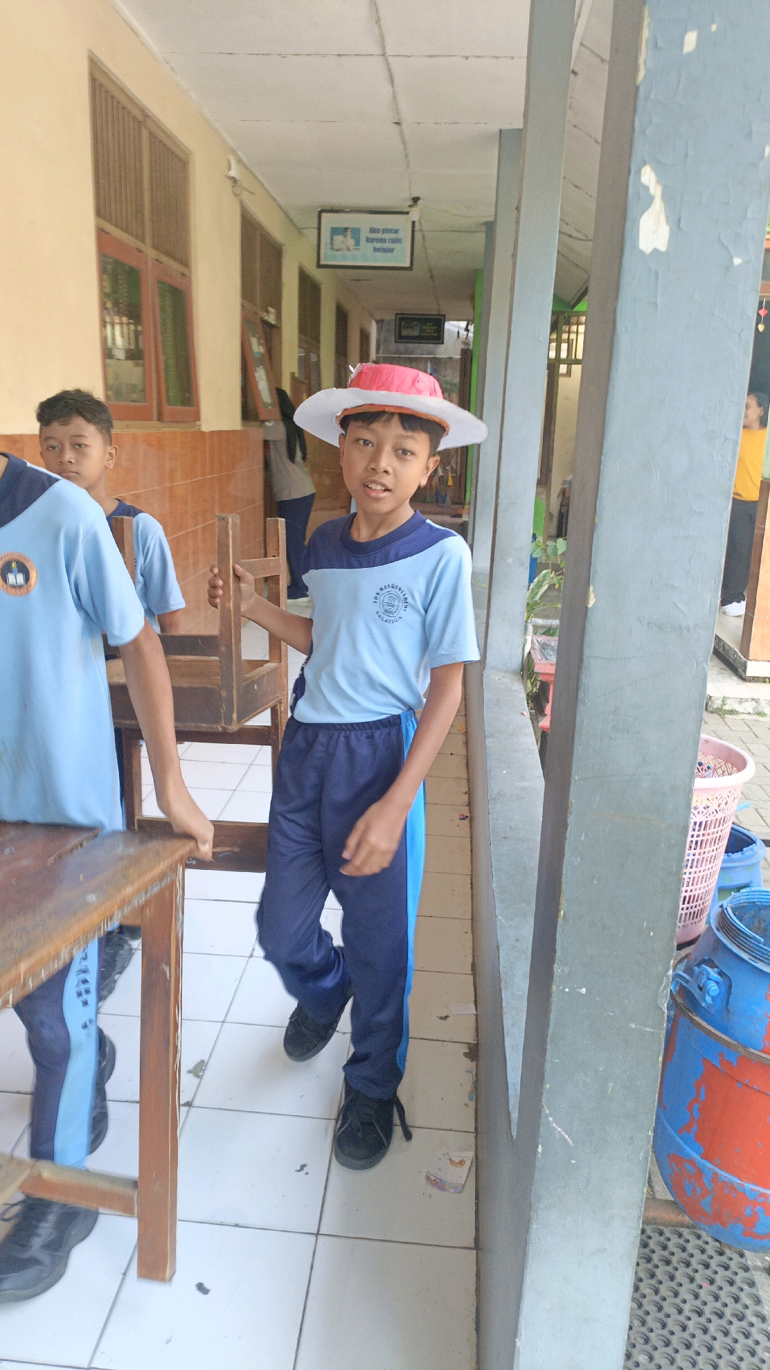 In a school hallway in Salatiga, Indonesia, a young boy wearing a light blue shirt, dark blue pants, and a handmade white and red hat is posing for the camera. He stands on a tiled floor, with a wall to his right and other students and a classroom in the background. A wooden chair is partially visible in his hand. The hallway appears to be an outdoor or semi-outdoor space, with natural light filtering in. The atmosphere is casual and appears to be a school event.