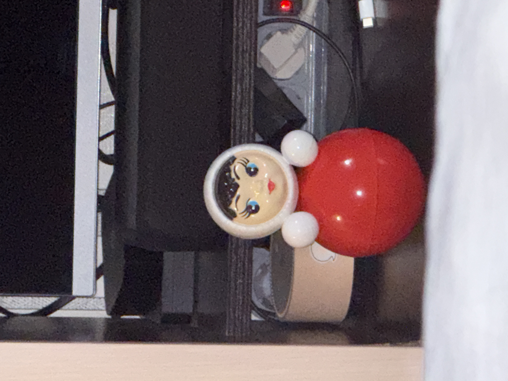 A close-up shot reveals a roly-poly toy, featuring a white face with painted facial features and a red, spherical body. The toy is situated in an indoor environment, nestled amidst various electronic devices and power cords. The surrounding area contains a black computer tower, cables, and an outlet surge protector. The toy rests on a light-colored surface, presumably a shelf or tabletop. The scene gives the impression of being a cluttered workspace. The location in the city of Unknown, Unknown is not known.