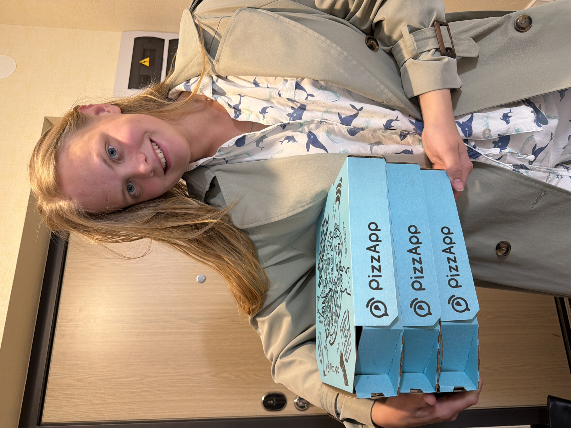 A young woman is holding three light blue pizza boxes. The boxes have "PizzApp" printed on them in black. The woman is wearing a light gray trench coat over a light-colored shirt with a whale pattern. She is smiling at the camera and has blonde hair and blue eyes. The background appears to be an interior setting with a light wood-paneled door and cream-colored walls. A black sign with a yellow hazard symbol is visible in the upper left corner. It is impossible to confirm the exact location in the city of Izhevsk, Russia based solely on the visual information provided.