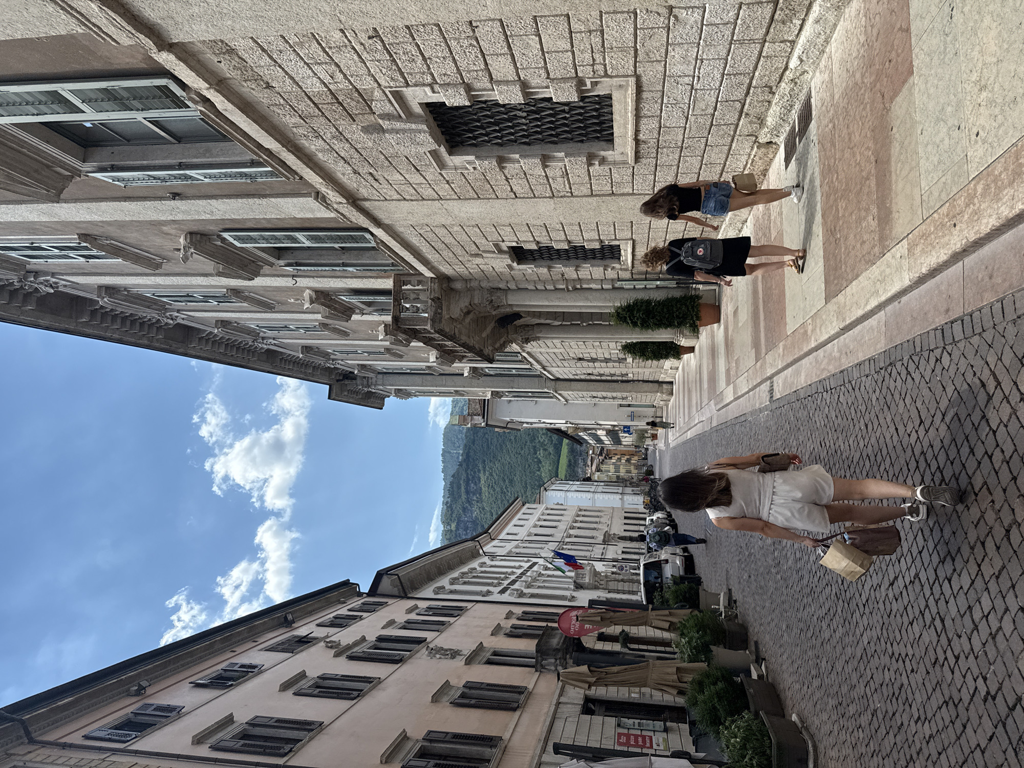 The image captures a street scene in Trento, Italy, specifically on Via Torre Vanga. On the right side of the street, a woman in a light-colored outfit walks away from the camera, carrying two shopping bags. She is wearing a sleeveless top and shorts, and white sneakers. To her right, two other women walk in the same direction. The woman closer to the camera has curly brown hair and is wearing a black dress and a backpack. The woman next to her has long dark hair and is wearing a black t-shirt and denim shorts, carrying a paper bag.
The street is paved with cobblestones. Buildings line both sides of the street, featuring Italian architectural styles with shuttered windows. On the left, a building with a pinkish facade has several shops with closed umbrellas. Further down the street, cars are parked, and more buildings with balconies are visible, leading towards a backdrop of lush green mountains under a partly cloudy blue sky. The overall impression is of a sunny day in a historic European city.