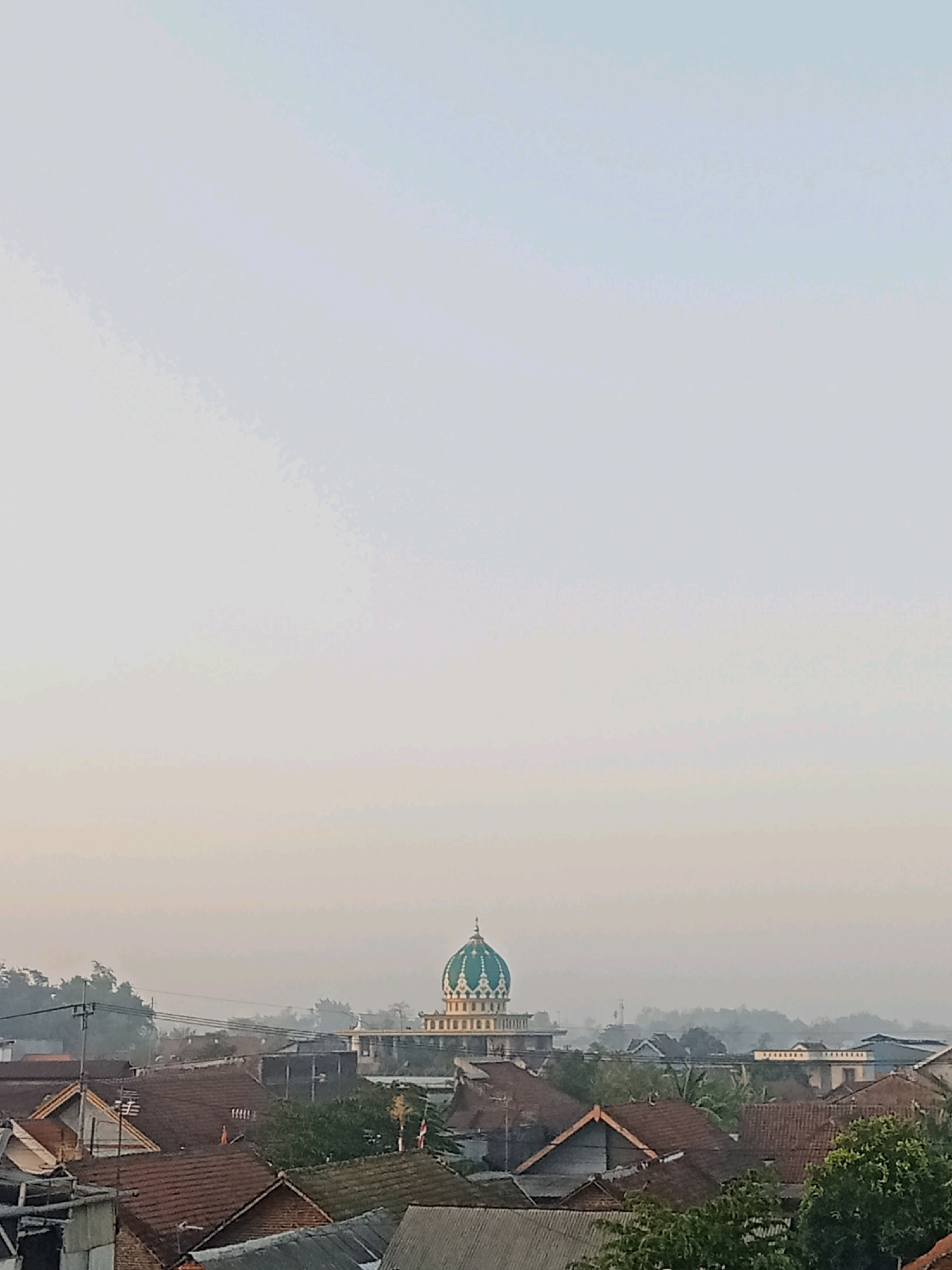 The image captures a hazy, overcast sky over a collection of rooftops in the city of Keplaksari, Indonesia. In the mid-ground, a prominent mosque stands out with its distinctive green dome adorned with white architectural details. Surrounding the mosque are numerous residential buildings with terracotta tiled roofs, some showing signs of age. Trees and power lines are visible interspersed among the structures, indicating a moderately developed urban or semi-urban environment. The overall atmosphere suggests an early morning or late afternoon due to the soft, diffused light and the presence of mist or fog. No people are visible in the frame.