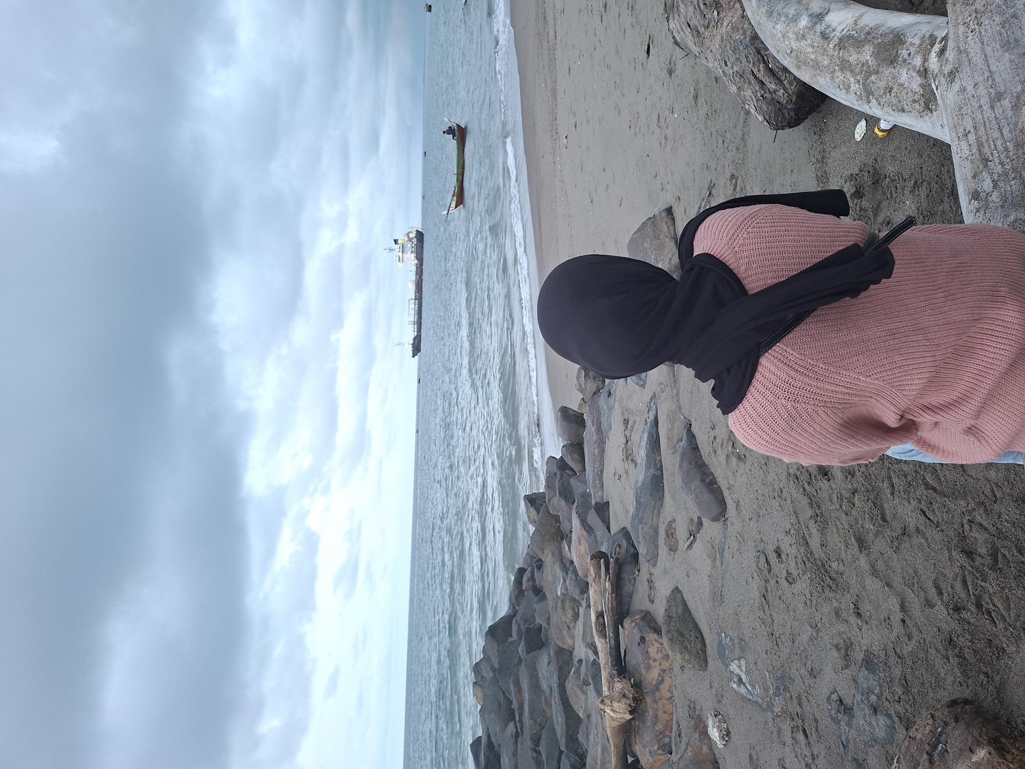 A woman wearing a black headscarf and a pink sweater is sitting on a beach. The scene is at a beach in Lhokseumawe, Indonesia. She is situated near a rocky breakwater with several large rocks lying on the sand. The beach stretches out along the coast, and there are two ships visible on the water in the distance. One is a larger cargo ship, and the other is a smaller boat. The sky is overcast with grey clouds. Large tree trunks are also lying nearby on the sand.