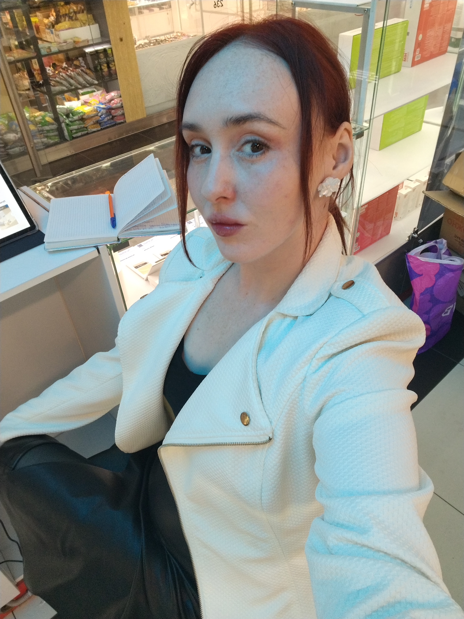 A woman is taking a selfie inside what appears to be a retail establishment. The woman has reddish-brown hair, fair skin, and is wearing a white, textured motorcycle-style jacket over a black top. She is also wearing earrings that resemble white flowers. She is seated, and what appears to be black leather pants or a skirt can be seen.
Behind her, there's a counter with a notebook and pen. There are shelves with packaged goods, possibly food or other retail items, visible in the background. Other items such as packaged cosmetics and a shopping bag can be seen on the shelves behind her. The store is located in Izhevsk, Russia, based on Cyrillic characters written on one of the packages in the background.