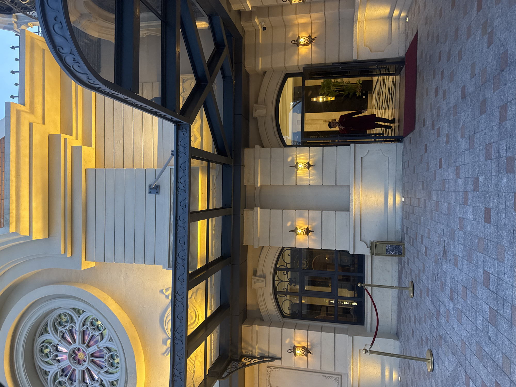 The image depicts the entrance of a building with a grand, ornate facade in Nha Trang, Vietnam. The architecture features classical elements, including a large rose window with stained glass on the upper level and decorative stonework. The building is illuminated by warm, yellow lighting, highlighting its textured walls and layered architectural details.
A modern glass and steel canopy extends over the entrance, providing shelter. Beneath the canopy, a person dressed in a maroon uniform, possibly a hotel staff member or security guard, is walking out of the building through double doors. The person is holding something to their ear, suggesting they are on the phone. The entrance area has a patterned tiled floor, and a red carpet is laid out before the doors.
Flanking the entrance are tall, stone columns and classical-style lanterns that emit a soft glow. The area is further enhanced by ground-level lighting and a velvet rope barrier supported by golden stanchions, indicating a controlled access point or a designated pathway. The overall impression is one of elegance and formality, typical of a high-end establishment.