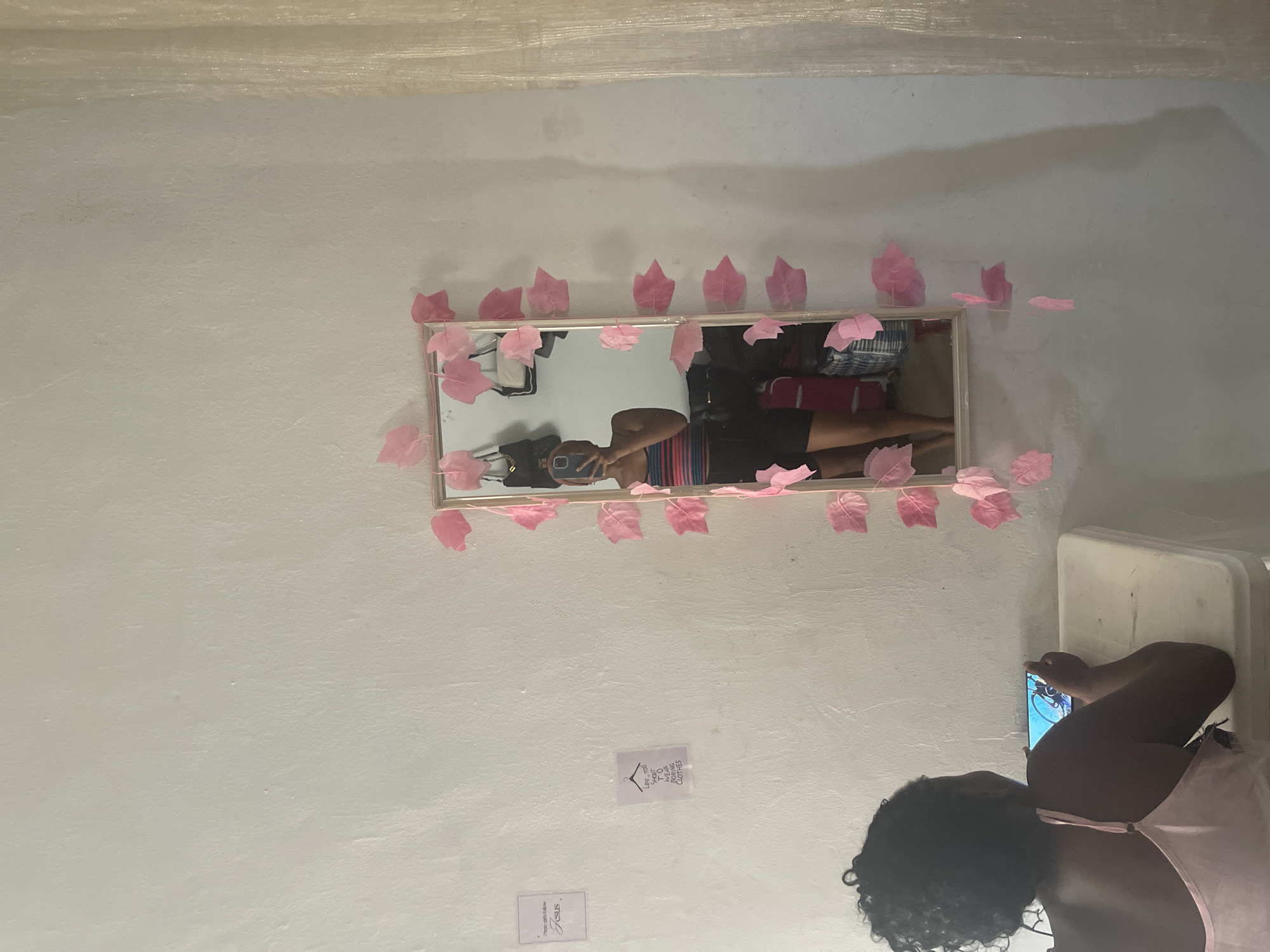 A person is taking a picture of themselves in a mirror. The mirror is decorated with a string of pink leaves. The person is wearing a pink tank top. In the reflection, the person is holding a phone. The wall behind the mirror is white. There are two small signs on the wall below the mirror. One sign has a drawing of a hanger and text that reads "LET YOU LOVE TO WEAR BETTER CLOTHES". The other sign has text that reads "Please tell them to follow Jesus". The scene appears to be indoors, possibly a bedroom or dressing area. The overall lighting suggests it is daytime.