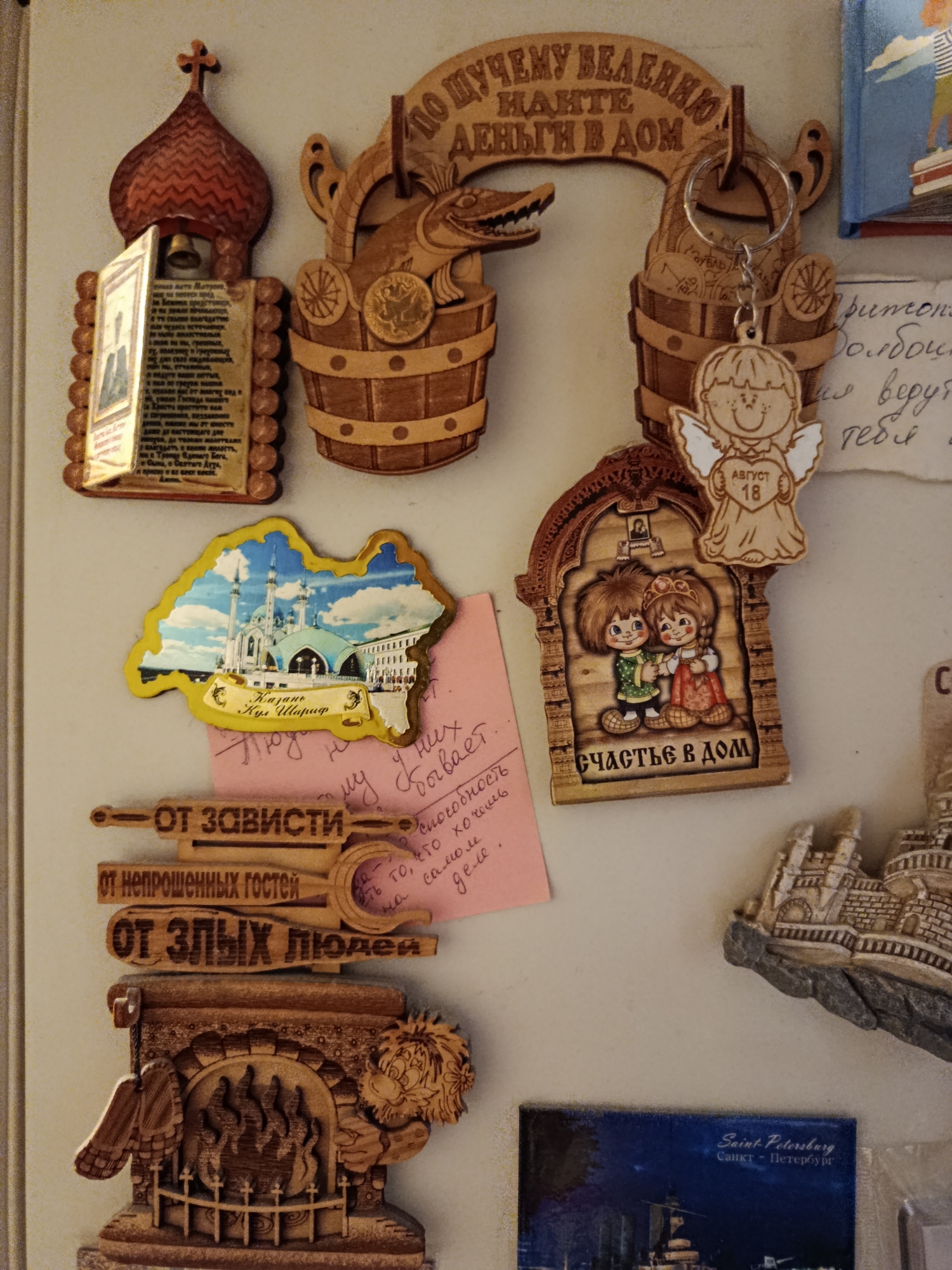 The image displays a collection of wooden souvenirs and a postcard attached to a refrigerator. The souvenirs feature religious and folkloric motifs, common in Russian culture. One prominent souvenir is a wooden icon of a church bell tower with a depiction of a saint inside. Another is a wooden bucket filled with coins and a small wooden angel keychain with the inscription "АВГУСТ 18" (August 18). There are also three horizontally oriented wooden plaques with inscriptions, seemingly protective charms: "ОТ ЗАВИСТИ" (From envy), "ОТ НЕПРОШЕННЫХ ГОСТЕЙ" (From uninvited guests), and "ОТ ЗЛЫХ ЛЮДЕЙ" (From evil people). Below these is a more elaborate wooden carving of a fireplace with a lion-like figure on the right. A postcard depicting the Kazan Kremlin in Kazan, Russia, is also visible. Another souvenir shows two children holding hands within an arched frame with the inscription "СЧАСТЬЕ В ДОМ" (Happiness in the home). The background consists of a cream-colored refrigerator door. There is no indication of the city of Saratov, Russia, in this image.