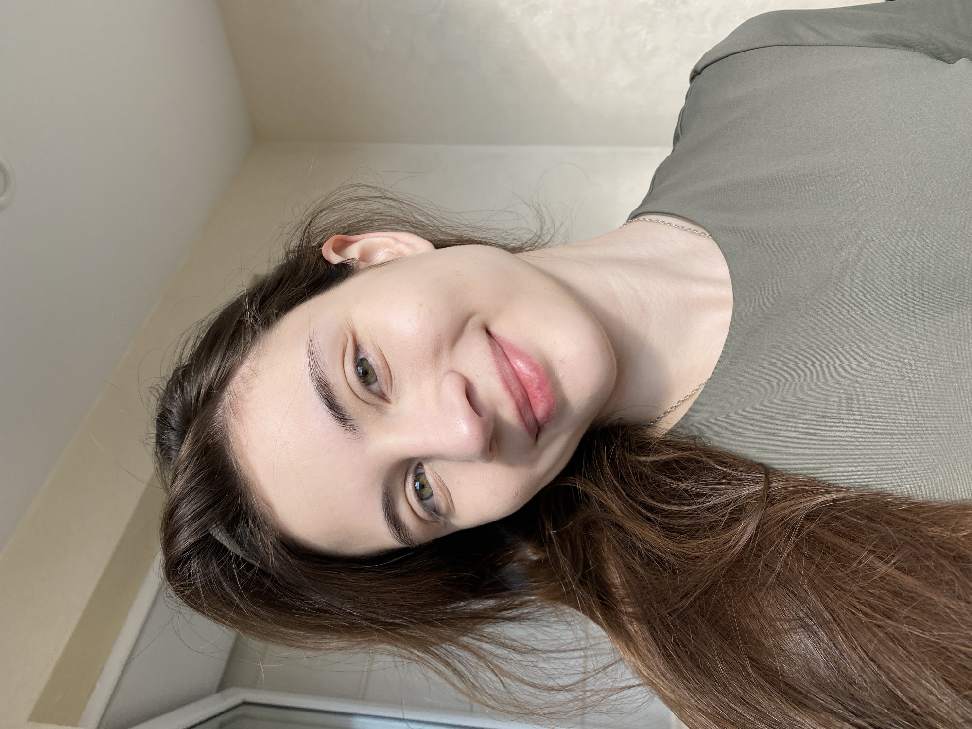 A portrait of a young woman is captured indoors in Votkinsk, Russia. The image, originally rotated sideways, shows her looking gently upwards, with her head softly tilted. She has fair skin, natural dark eyebrows, and striking green eyes that engage the viewer. Her lips are a soft pink, forming a subtle, sweet smile that conveys a sense of calm and approachability.
Her long, wavy hair is a rich dark brown with visible lighter, auburn-toned streaks, flowing gracefully over her left shoulder and chest. She is dressed in a plain, round-neck top of an olive green or dark sage color. A delicate silver chain necklace is visible around her neck, resting against her skin.
The background consists of a simple, light-colored wall, likely off-white or light beige, indicating a clean and bright indoor setting. A hint of a window frame or similar architectural detail is visible in the lower left corner of the frame (when viewed correctly). The lighting is soft and even, suggesting either diffused natural light from a window or gentle indoor lighting, implying a daytime setting. There is no visible text within the image. The overall mood is one of natural grace and quiet contentment.