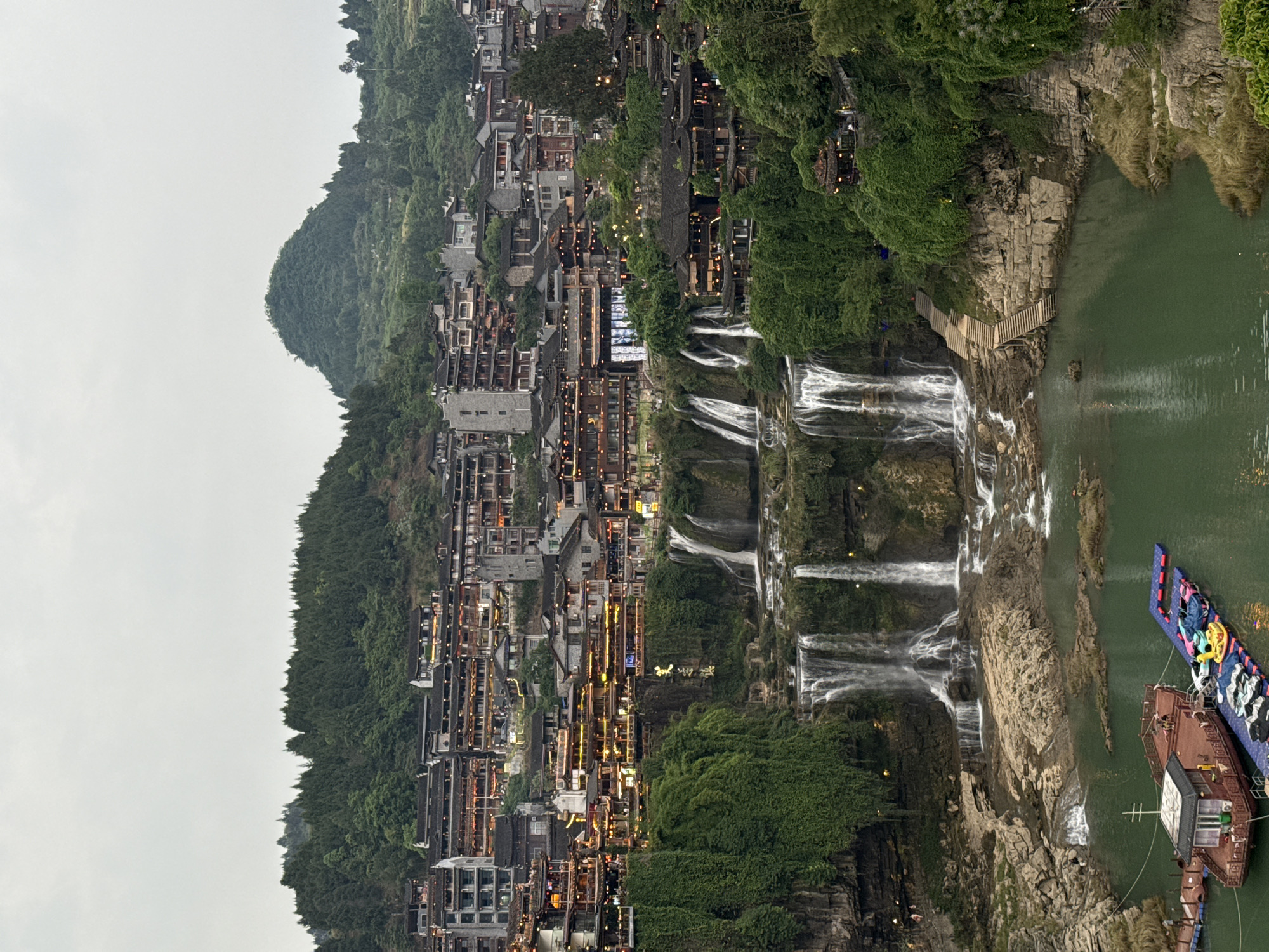 The image depicts a hillside village integrated with a cascading waterfall, likely taken in the city of Furong, China. The village is constructed along the slope with buildings of varying heights and traditional architecture, some illuminated with warm lights. The waterfall flows through the village, interspersed with greenery and rocky outcrops. In the lower right corner, two boats are moored in the water. The sky is overcast and grey.