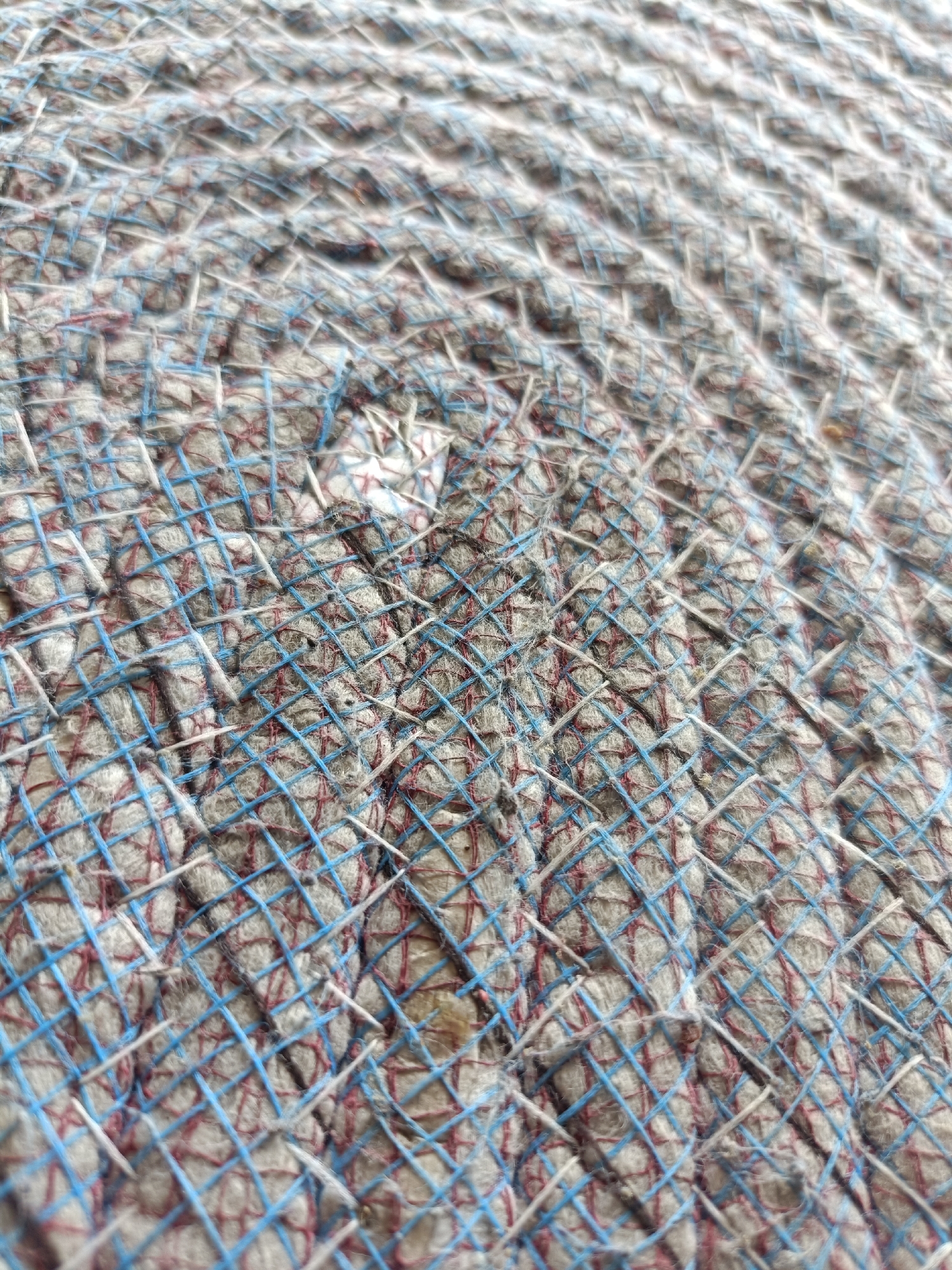 The image shows a close-up, abstract view of a woven textile. The weave is circular, forming a spiral pattern, and appears to be made of coarse, grey fibers interlaced with thin blue and red threads. The threads create a diamond-like grid over the textured surface. There are no visible people, specific objects, or environmental details that would indicate a location in Kediri City, Indonesia, or any other specific place. The image focuses solely on the texture and pattern of the fabric.