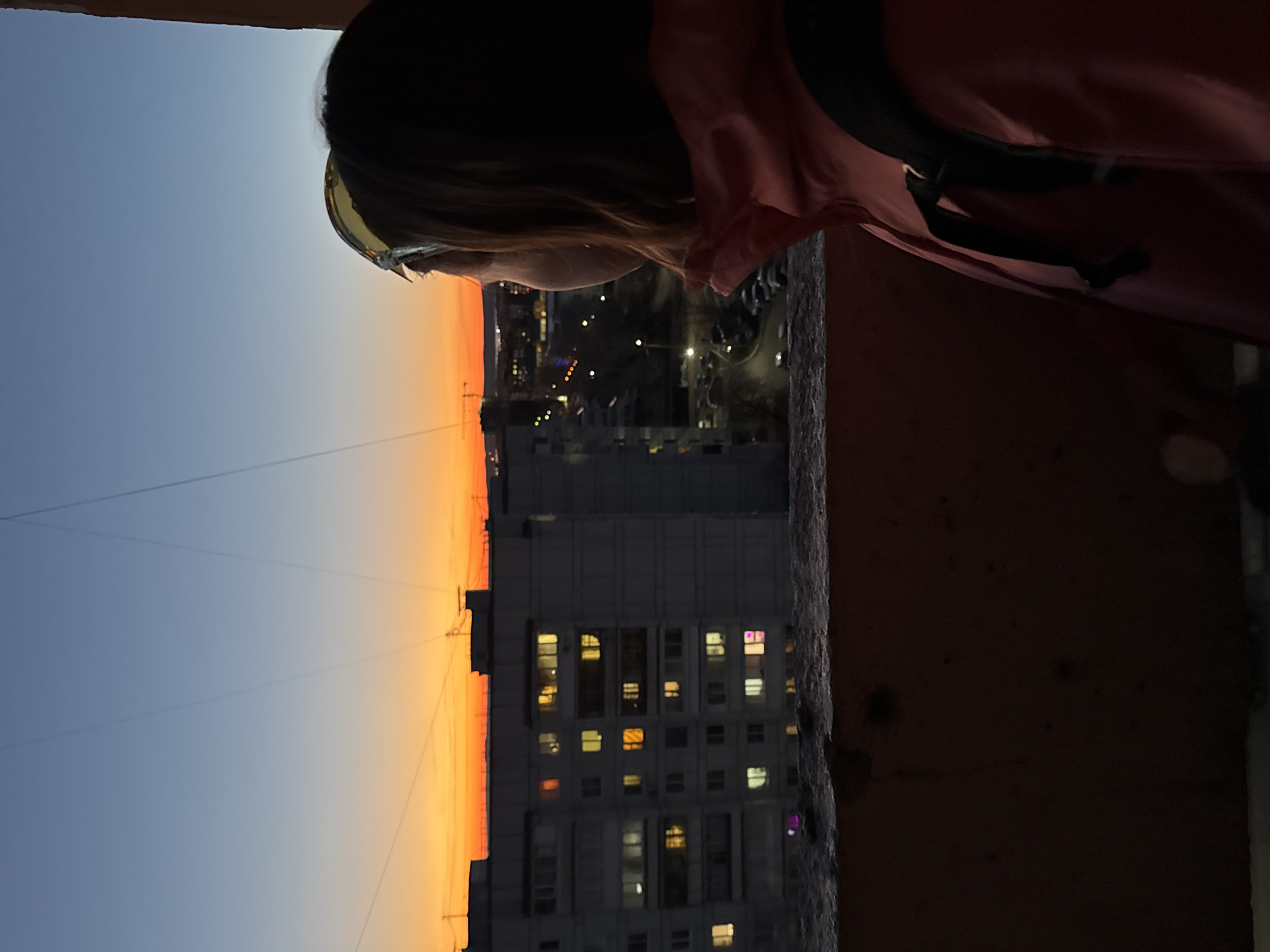 A person with dark hair and wearing sunglasses and a reddish jacket with a black strap stands at the edge of a balcony overlooking a cityscape at dusk. A tall, grey apartment building is visible in the center of the view, with lights on in some of the windows. In the distance, blurred city lights are seen. The sky transitions from orange near the horizon to blue higher up. Thin cables stretch across the upper part of the frame. The balcony edge is rough and textured. The location is Chaikovsky, Russia.
