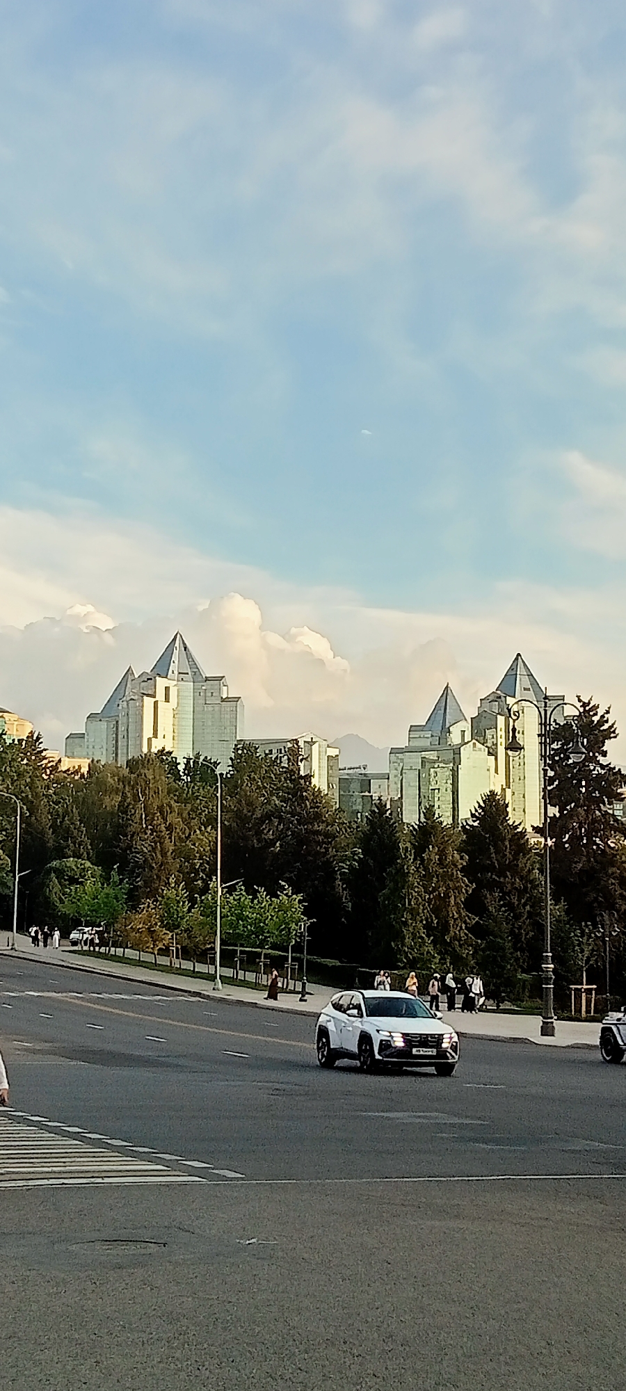 The image depicts a cityscape in Almaty, Kazakhstan, featuring the distinctive architecture of the Presidential Palace and surrounding buildings. In the foreground, a white SUV is captured driving on a paved road, with a pedestrian crossing visible on the left. A line of trees separates the road from a sidewalk where several pedestrians are walking. Behind the trees, the imposing, angular structures of the Presidential Palace complex, with their signature pyramidal roofs, dominate the midground. The sky above is a mix of blue and scattered clouds. The overall scene suggests a daytime urban environment with a backdrop of notable architectural landmarks.