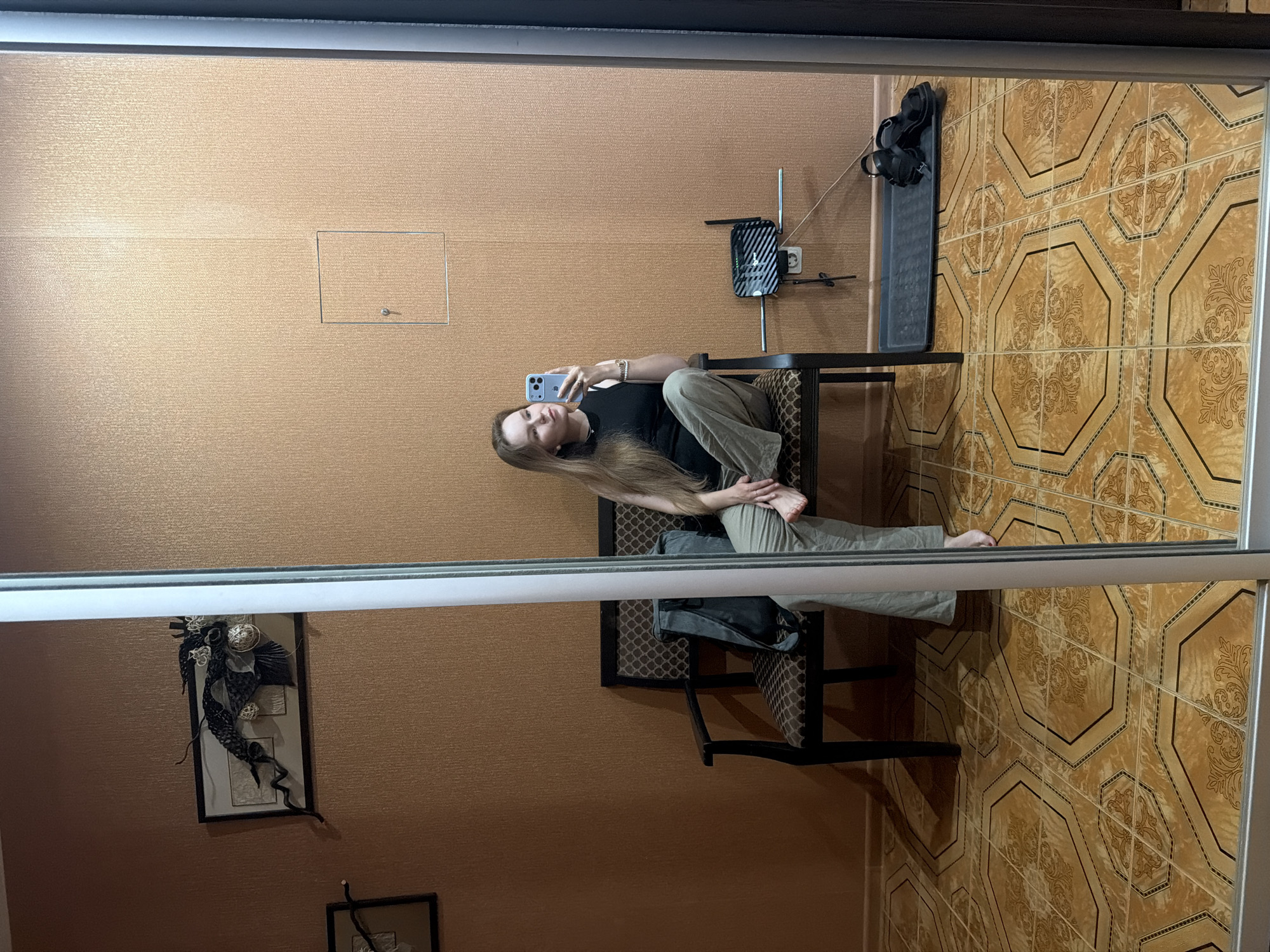In Kazan, Russia, a young woman with long blonde hair is seen in a mirrored reflection, sitting on a dark-colored bench with a patterned cushion. She is wearing a black sleeveless top and loose, light-colored pants. She holds a light blue smartphone in her right hand, taking a selfie.
Behind her, the wall is covered in a textured, light brown wallpaper. On the wall, a framed piece of dark, abstract wall art is mounted. To the right of the woman, a shelf is attached to the wall, holding a black router with antennas and a small electrical outlet. Below this shelf, a black tray contains a pair of black sandals. The floor is covered in large, octagonal tiles with a decorative pattern in shades of yellow and brown. The image is captured in a room with a reflective surface, likely a large mirror or a mirrored closet door.