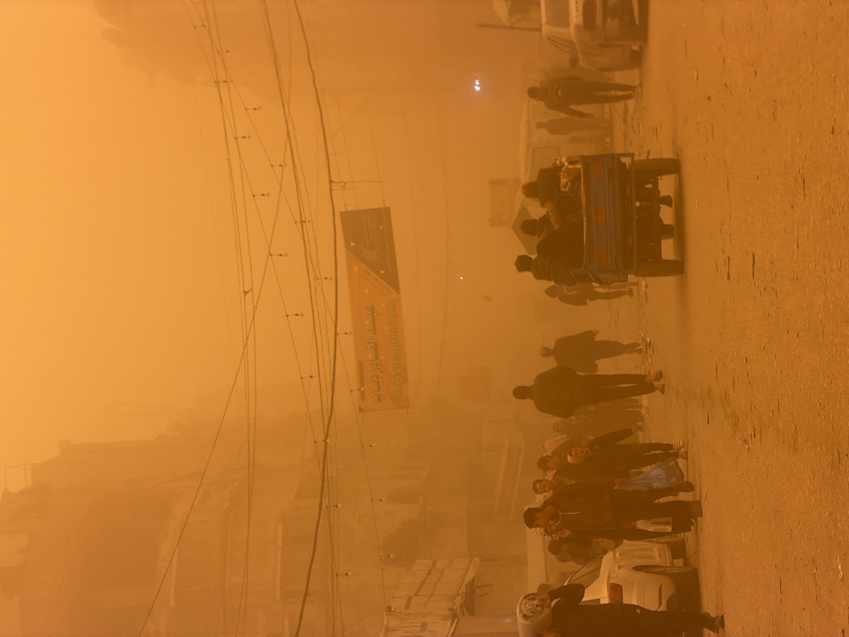 The image captures a street scene in the city of Unknown, Unknown, during an severe orange-brown dust storm that significantly reduces visibility. The entire environment is permeated by fine particulate matter, giving the scene a monochromatic, low-light appearance.
Approximately 15-20 individuals, predominantly adult males, are observed walking on the street and adjacent areas. On the far left, a man is carrying a white gas cylinder on his right shoulder. Other pedestrians appear to be moving with no discernible interactions, some carrying bags.
In the mid-ground, a dark-colored three-wheeled motorized vehicle, seen from the rear, travels down the street with 3-4 passengers visible in its cargo bed. On the left side of the street, a white sedan is partially visible, either parked or moving slowly. Another light-colored vehicle is partially obscured by dust on the right side further down the street.
Overhead, multiple utility lines with strung light bulbs traverse the street. A large banner displaying Arabic script is suspended above the street in the mid-distance. Buildings line both sides of the thoroughfare, their details largely obscured by the pervasive dust. The ground surface appears to be a paved or dirt road, covered with a layer of dust.