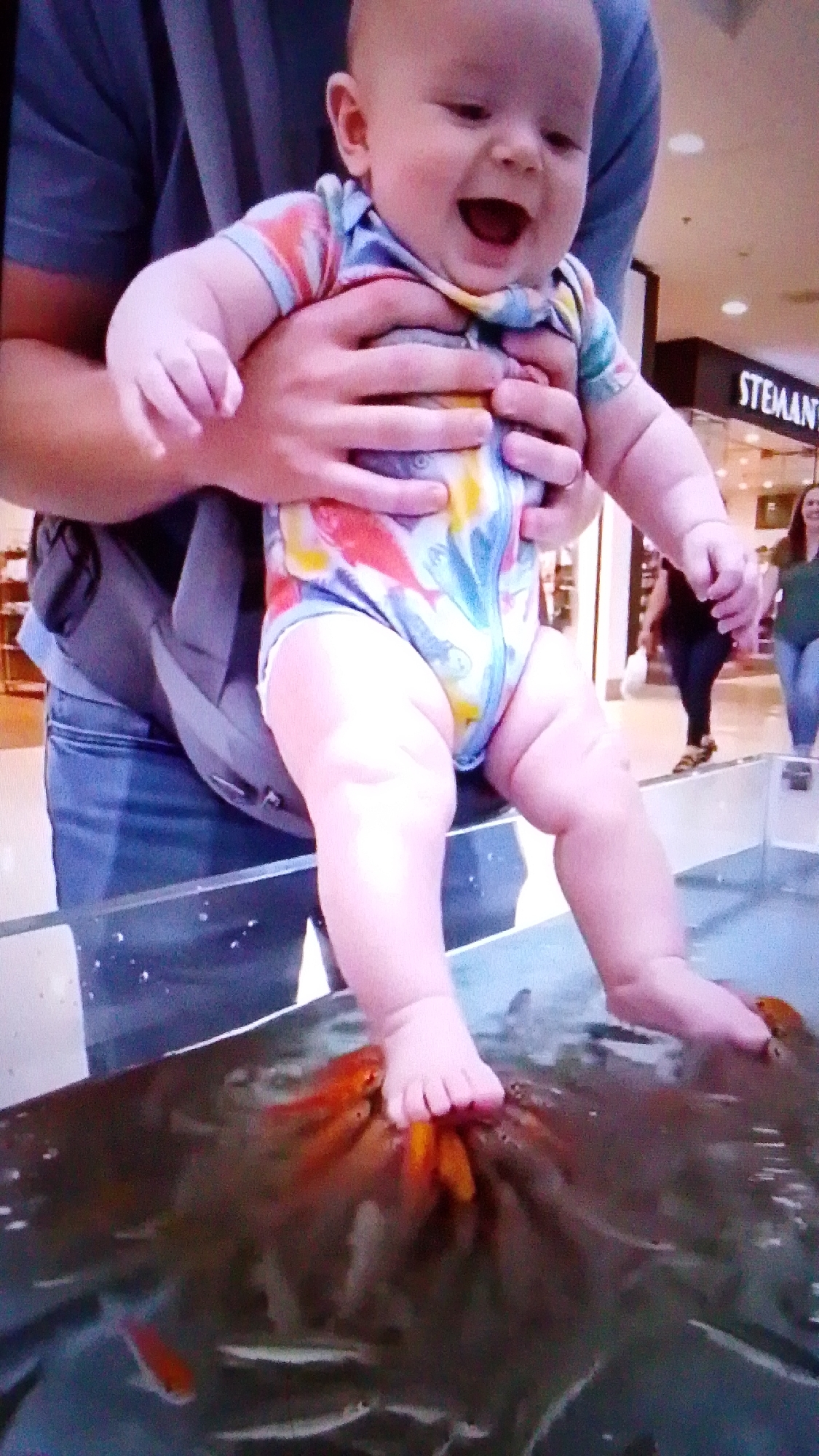 A baby wearing a colorful bodysuit is held by an adult in what appears to be a public space. The baby is leaning over a tank filled with water and small fish, with their feet submerged. The baby's head is turned to the side and they are laughing, with their mouth wide open. The background shows a store with a sign that says "STEMANT". There are other people in the background, including a woman in a green shirt and another woman in a black shirt and jeans. The lighting is bright, and the image is slightly pixelated. The overall scene suggests a playful and curious interaction between the baby and the fish.