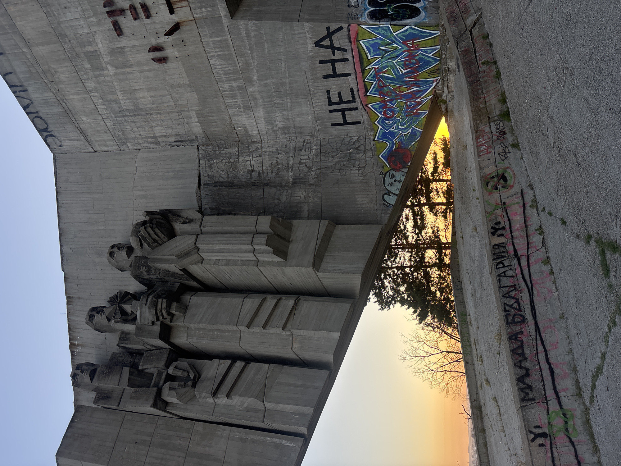 The image depicts the Buzludzha Monument, a brutalist architectural landmark in Bulgaria, located on Buzludzha Peak, approximately 20 kilometers from the city of Kazanlak. The monument is made of concrete and features large, imposing sculptures of figures. Graffiti adorns the concrete surfaces. The foreground shows a sloped concrete area with scattered grass and more graffiti. In the background, a tree-lined landscape is visible against a sunset sky with hues of orange and yellow. The overall scene suggests abandonment and decay, with the graffiti adding a layer of urban decay to the monument.