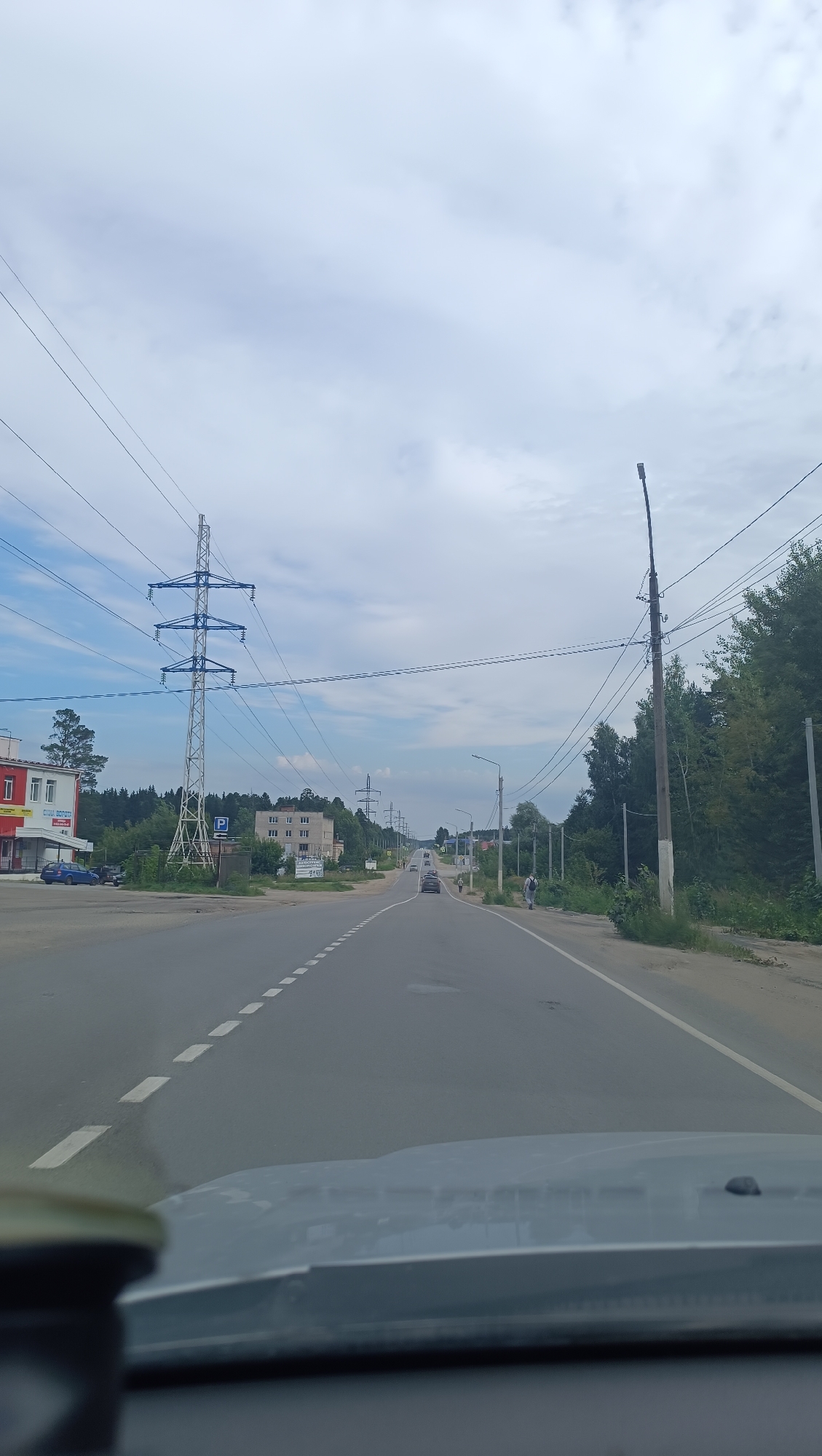 The image depicts a view from a vehicle traveling on a two-lane asphalt road in Chaikovsky, Russia. The road is bordered by a grassy shoulder and trees on the right, and a sidewalk and buildings on the left. Overhead power lines traverse the scene, supported by tall metal towers and wooden poles. Several cars are visible on the road, and a few pedestrians are walking on the sidewalk. The sky is overcast with clouds. On the left side of the road, there is a red and white building with a sign that reads "СТРОЙ МАТЕРИАЛЫ" (building materials). Further down the road, other buildings and more power infrastructure are visible. The overall impression is of a semi-urban or suburban setting.