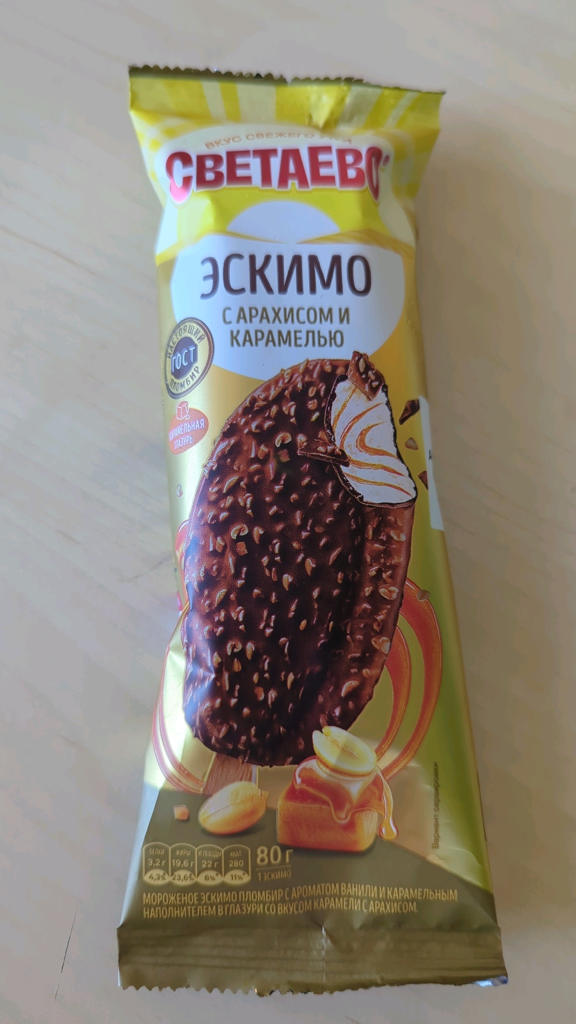 The image shows a close-up, eye-level shot of a golden ice cream wrapper. The wrapper features a bold red logo at the top that reads "СВЕТАЕВО" (Svetaevo). Below the logo, the word "ЭСКИМО" (Eskimo) is written in large white letters. Further down, smaller text reads "С АРАХИСОМ И КАРАМЕЛЬЮ" (with peanuts and caramel). A prominent illustration of a chocolate-covered ice cream bar with nuts and a caramel swirl is the central focus. Nutritional information is displayed in a table at the bottom, with details such as protein, fat, carbohydrates, and calories. The bottom text describes the product as "МОРОЖЕНОЕ ЭСКИМО ПЛОМБИР С АРОМАТОМ ВАНИЛИ И КАРАМЕЛЬНЫМ НАПОЛНИТЕЛЕМ В ГЛАЗУРИ СО ВКУСОМ КАРАМЕЛИ С АРАХИСОМ" (Eskimo ice cream plombir with vanilla flavor and caramel filling in a glaze with caramel and peanut flavor). The background is a light-colored, wooden surface. The location of Большой Лог, Russia is not visually discernible from the image itself.