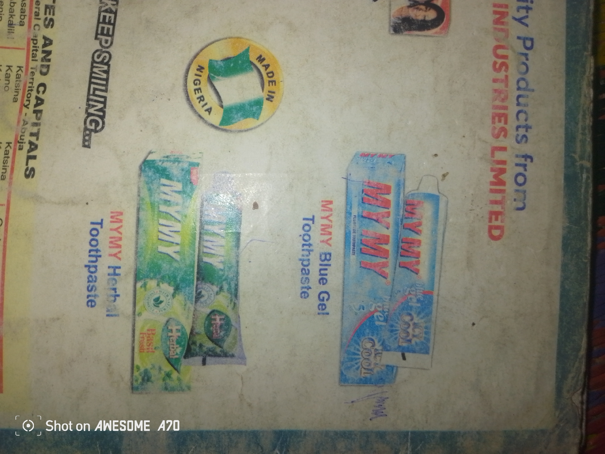 The image is a close-up, tilted, overhead shot of a light-colored, textured surface, possibly paper or a thin board, with several items placed on it. The surface is worn and shows signs of age and handling, with smudges and faint markings.
Prominently displayed are three tubes of "MYMY" brand toothpaste, arranged in a slightly diagonal line. The leftmost tube has a green and white label with text that appears to be in English. The middle tube is also green and white, with similar branding. The rightmost tube is blue and white, with the text "MYMY Blue Gel" and "Cool Cool" visible.
To the left of the toothpaste tubes, vertical text in a darker color reads "KES AND CAPITALS" and "KEEP SMILING." Further to the left, there are more lines of vertical text in red and black, some of which are partially obscured or difficult to read.
In the upper left quadrant of the image, there is a circular emblem with the words "MADE IN NIGERIA" and a depiction of the Nigerian flag. To the right of this, in the upper right quadrant, a small rectangular image shows the profile of a person.
Along the right edge of the image, vertical text in dark blue reads "ity Products from INDUSTRIES LIMITED."
At the bottom left corner of the image, there is a watermark that reads "Shot on AWESOME A70."
The overall impression is that of a collection of products and branding information, possibly a display or a sample board, laid out on a surface. The lighting is somewhat uneven, casting faint shadows.