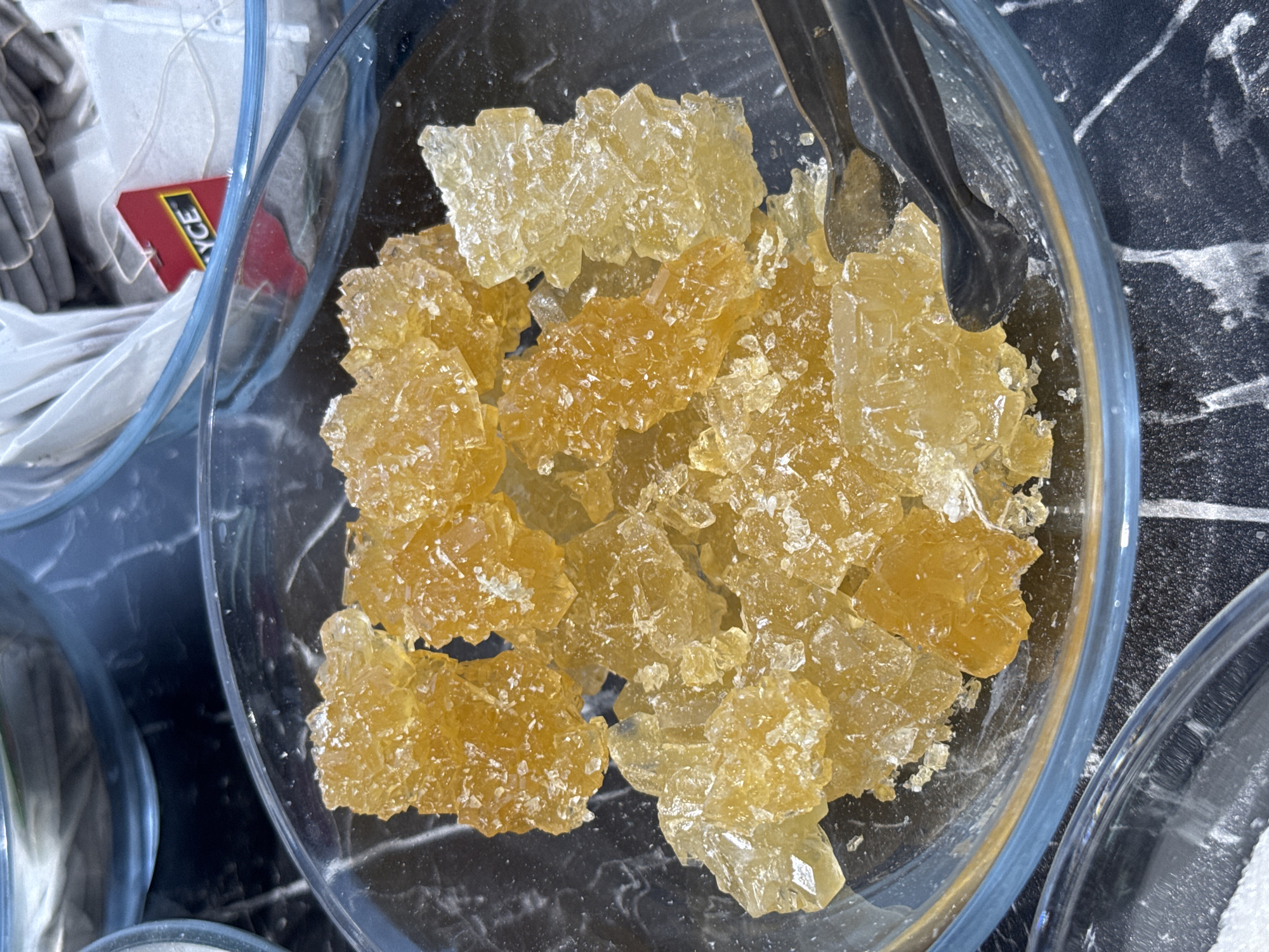 A high-angle shot depicts a glass bowl filled with amber-colored, crystallized sugar. A pair of black tongs rests in the bowl. Adjacent to the sugar bowl, a container holding tea bags is visible; a red tea tag with white lettering is partially visible. The background is a black surface with white marbling. The location is unspecified, but presumed to be in Tashkent, Uzbekistan.