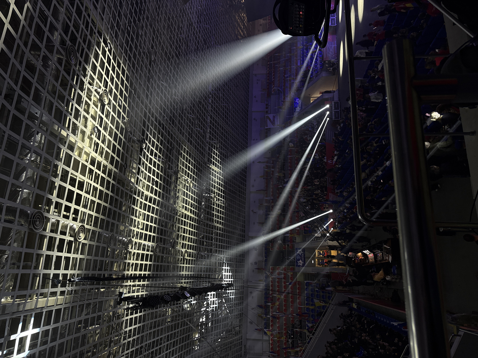 The image depicts an interior space, likely an arena or large hall, with a high ceiling featuring a grid-like structure. Spotlights illuminate the scene, projecting beams of light towards a crowd seated in tiered rows. The crowd appears dense, suggesting a well-attended event. The environment is dark, with the focus on the illuminated areas. Metal railings are visible, potentially from an upper level vantage point. Air vents are apparent on the grid-like structure.