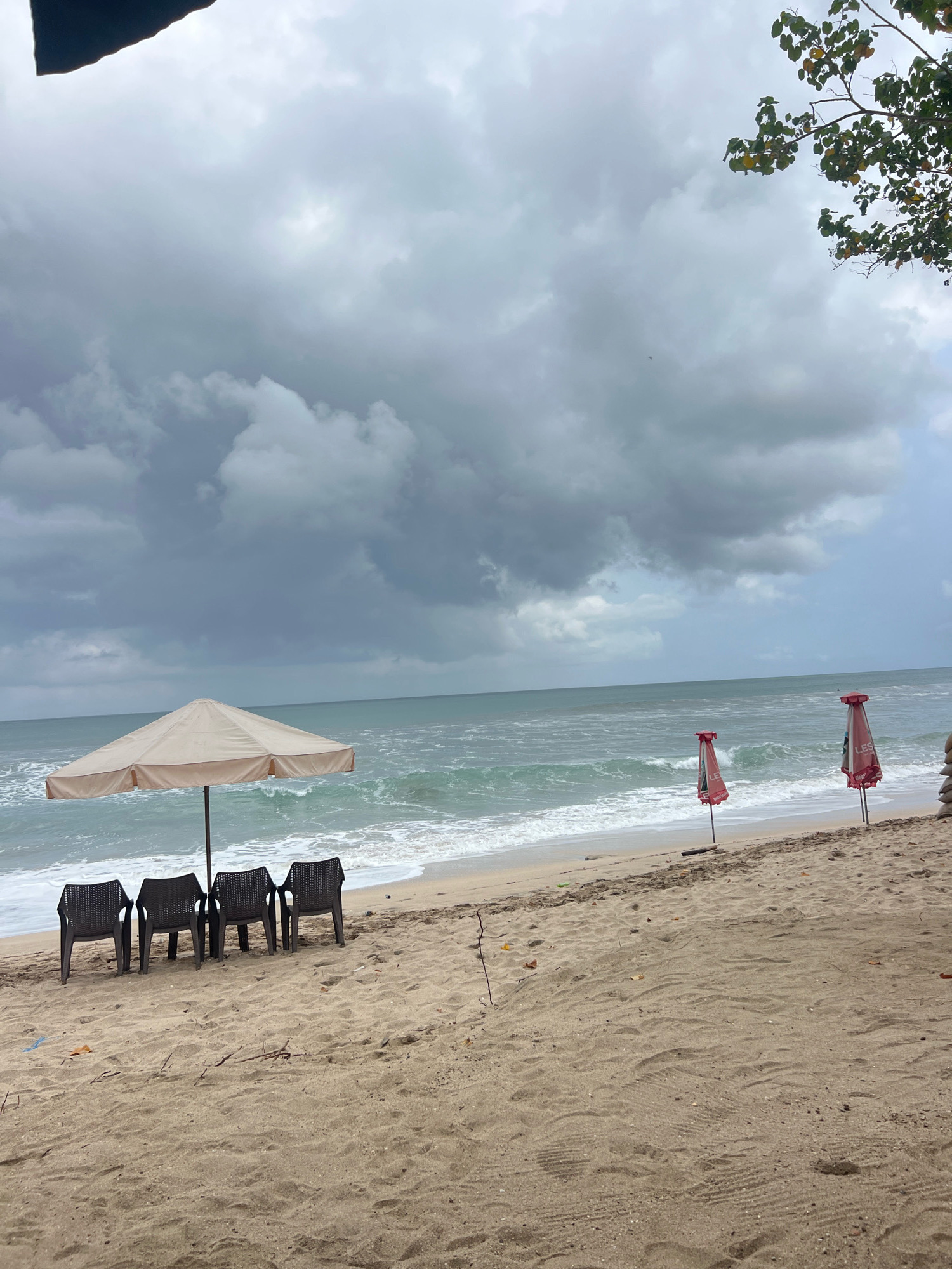 The image depicts a beach scene on a cloudy day. In the foreground, there is a sandy beach with several dark plastic chairs arranged under a large, light-colored umbrella. Beyond the chairs, the ocean stretches out, with moderate waves breaking on the shore. Further down the beach, two closed red umbrellas are planted in the sand. The sky is overcast with dark, heavy clouds, suggesting an impending rain or a storm. Foliage from a tree is visible in the upper right corner of the frame. The environment appears to be a beach resort or public beach area, potentially located in Kuta, Indonesia, known for its beaches and tourist amenities.