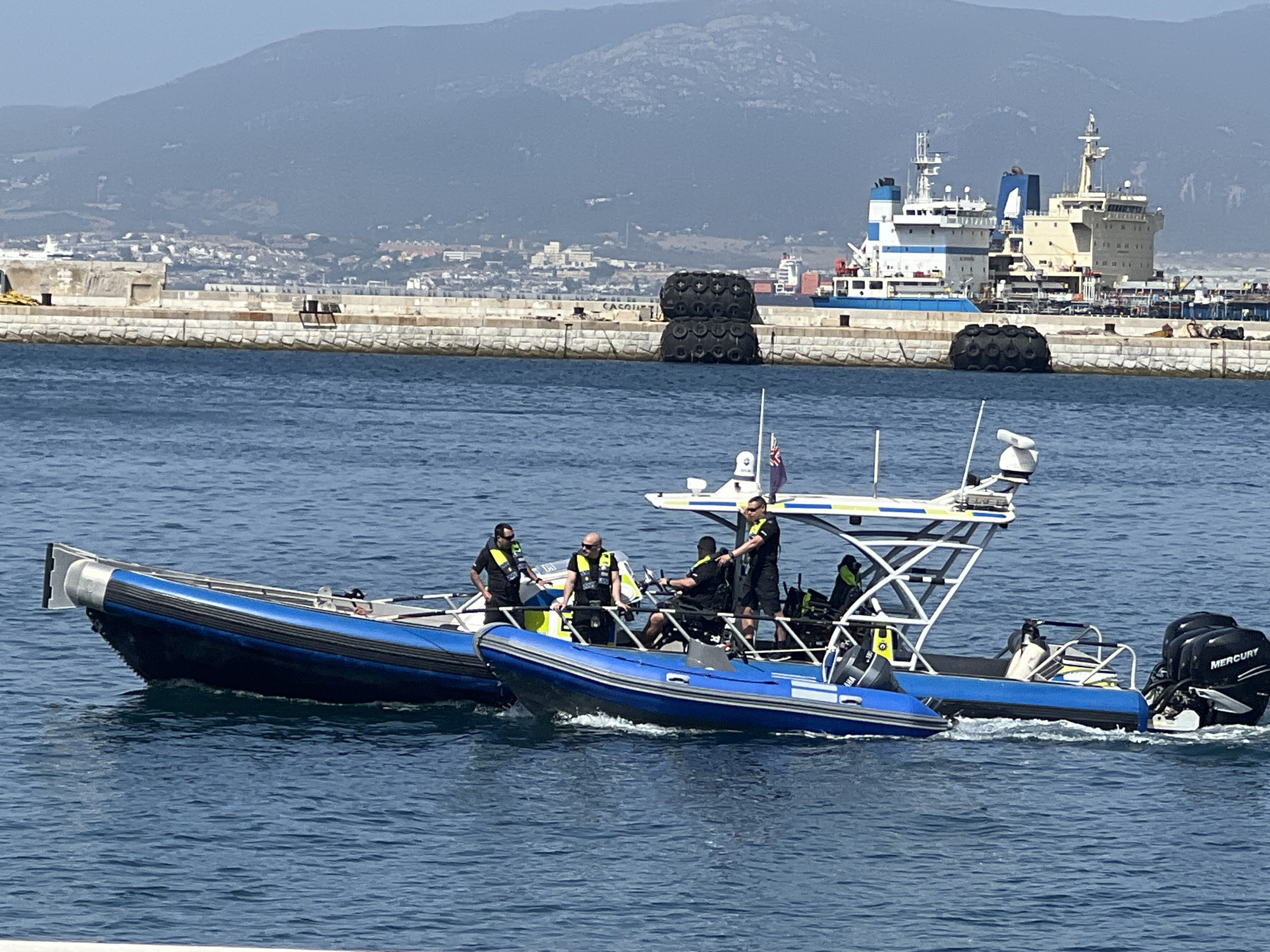 In the waters of Gibraltar, Gibraltar, two blue inflatable boats are underway. The larger boat in the foreground has a white canopy and is crewed by at least four individuals in dark uniforms and life vests. One person is steering, another is standing and looking forward, and two others are seated. The boat is powered by twin Mercury outboard motors. A smaller blue inflatable boat is to the left of the larger one, also with people on board. In the background, a stone quay is visible, with large black tires stacked on it. Beyond the quay, a large blue and white cargo ship is docked, and a mountainous landscape with scattered buildings forms the backdrop under a clear sky.