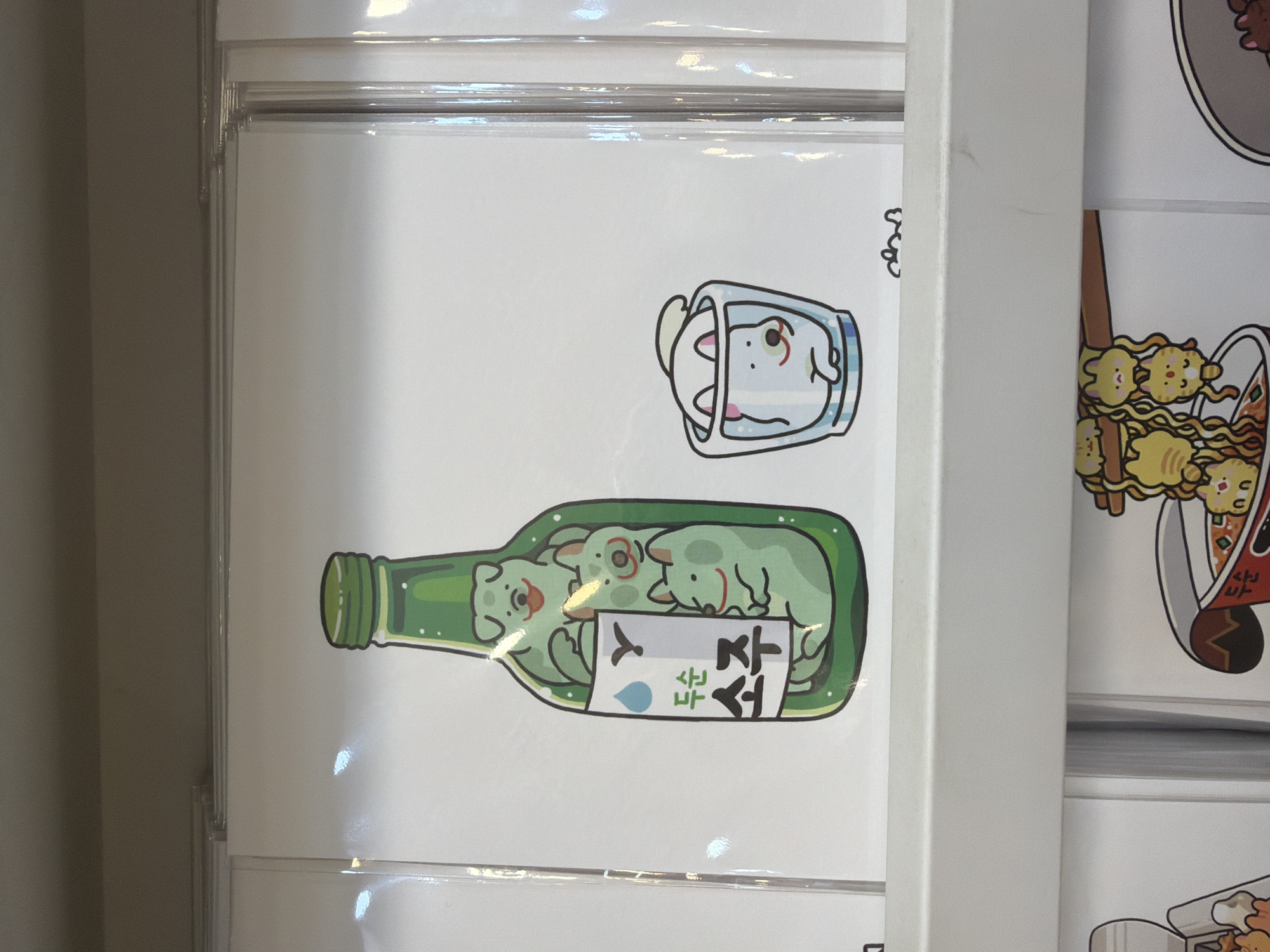 The image shows a display of merchandise, likely in a retail setting in Seoul, South Korea. The primary subject is a print featuring a cartoon illustration of green dogs inside a green soju bottle, with one white dog in a glass on the right. The soju bottle has a label with Korean text that appears to say "두순 소주" (Dusoon Soju). The artwork is presented in a clear plastic sleeve. Below this print, another row of merchandise is partially visible, including a depiction of cats hanging from noodles in a cup. The background is a plain, light-colored surface.