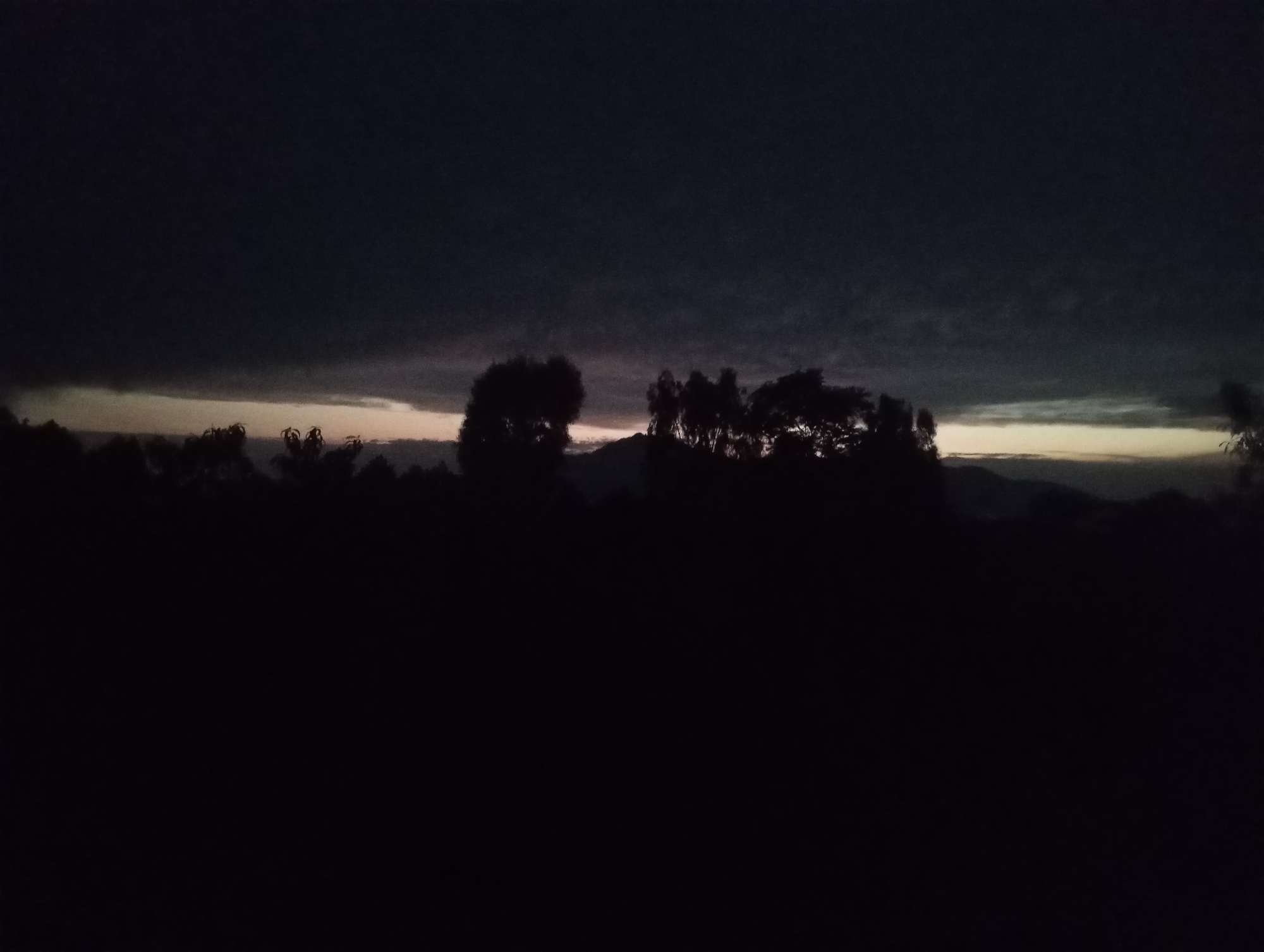 The image depicts a dark, silhouetted landscape, likely at dusk or dawn. The foreground is almost entirely black, obscuring any ground details. Above this dark expanse, a band of lighter sky is visible, with a few scattered clouds. The middle ground features the dark outlines of trees and possibly hills or mountains in the distance. The overall impression is one of low light and a natural environment. The scene is devoid of human presence or any man-made structures.