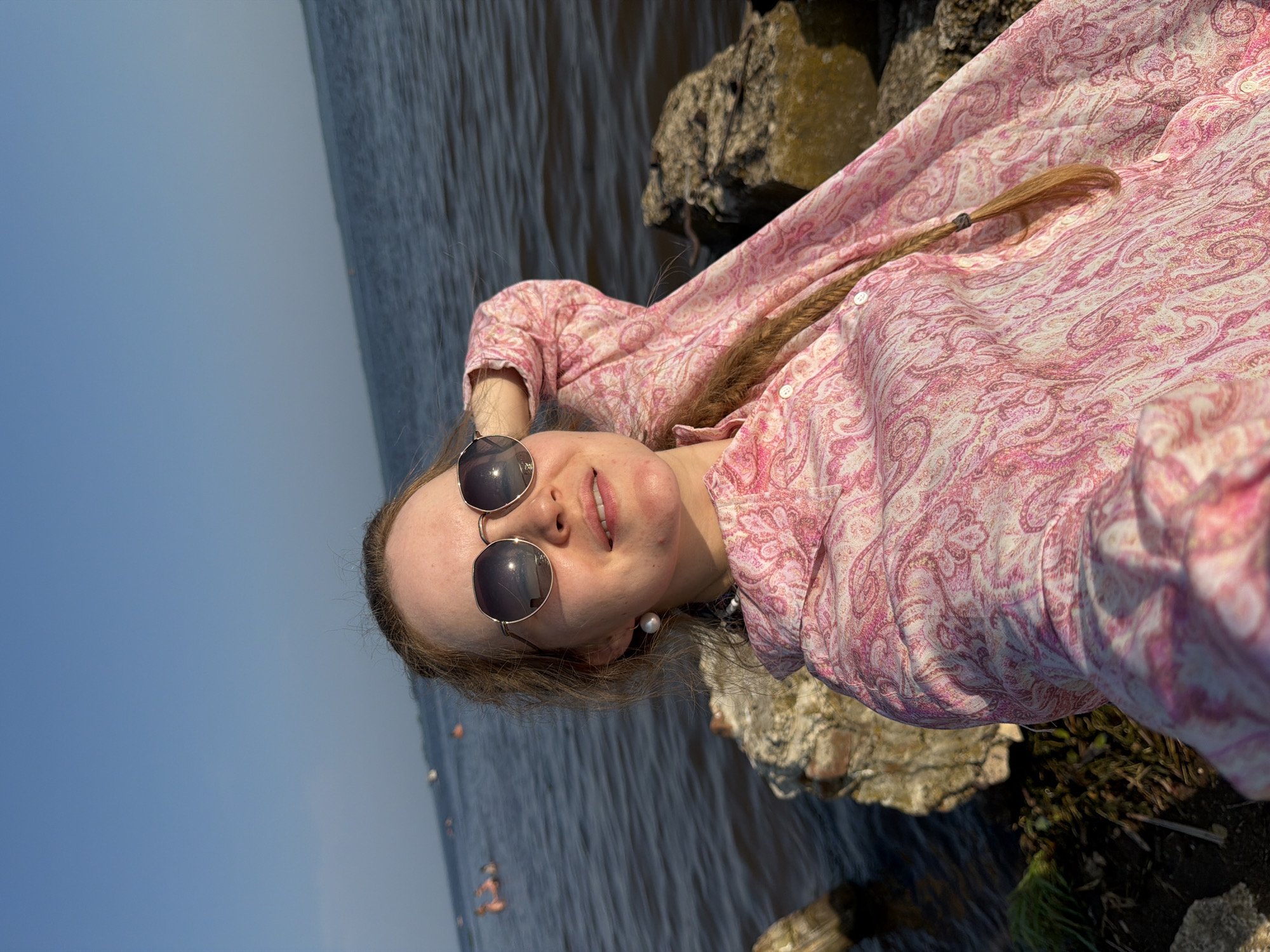 A young woman with blonde hair braided into a pigtail is taking a selfie by a body of water. She is wearing sunglasses and a pink paisley-print shirt. The woman is sitting on rocks by the water. In the background, there are people swimming in the water. The sky is clear and blue. The overall impression is one of a sunny day spent outdoors.