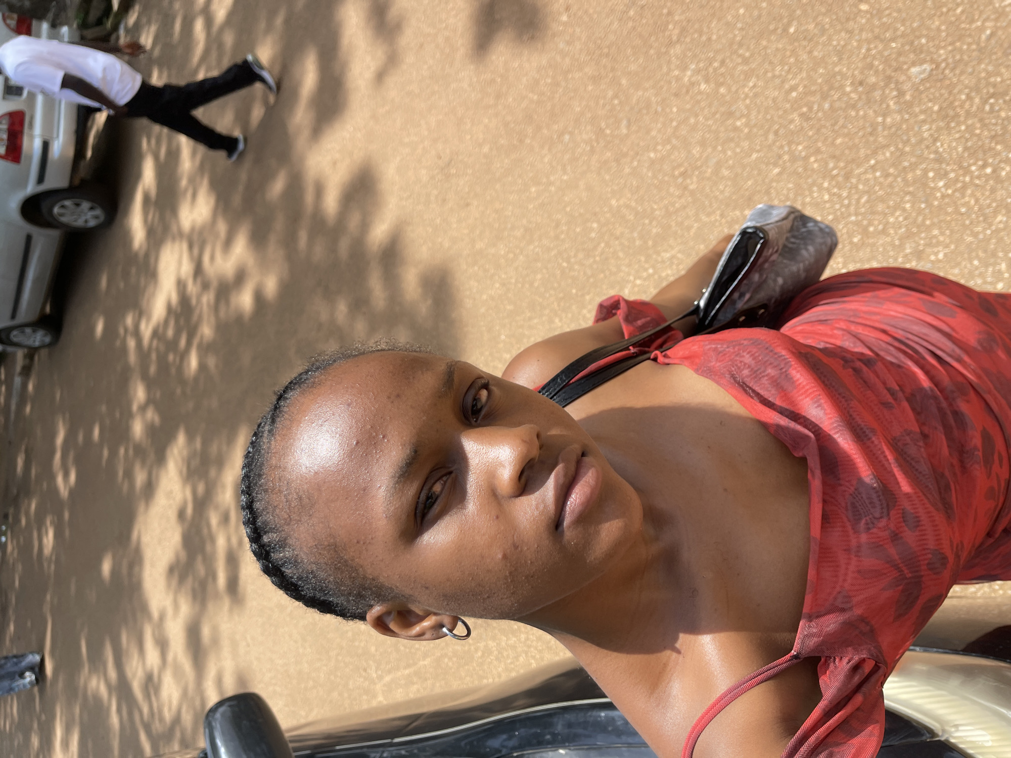 The image depicts a close-up, overhead shot of a young Black woman wearing a red, sheer, floral-patterned dress. She has her hair braided and is wearing a silver hoop earring in her left ear. She is holding a dark-colored purse with a patterned texture on her right side. The woman is positioned in front of a black car, and the background shows a sandy ground with scattered shadows from trees. In the distance, a silver SUV and a person wearing a white t-shirt and black pants can be seen. The overall setting appears to be an outdoor area, possibly a street or a pathway, with a light brown, sandy surface. The image does not provide specific information about the location in the city of Rafin Par, Nigeria.