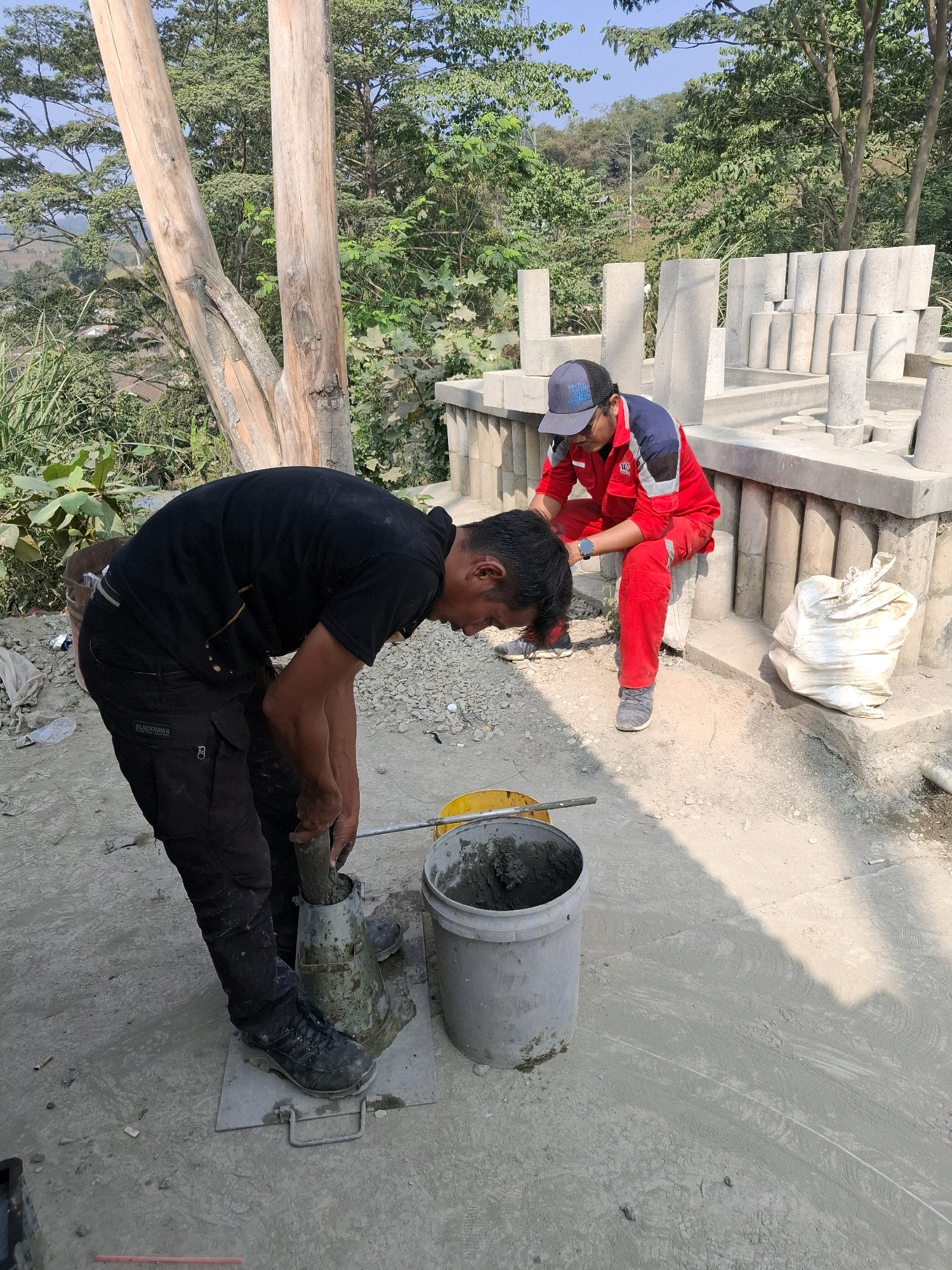 Two men are working outdoors on a construction site. The man in the foreground, wearing a black t-shirt and black pants, is bent over and using a metal rod to compact material into a cone-shaped mold. He is standing on a metal plate. To his right is a gray bucket containing concrete. In the background, a man wearing a red and blue uniform and a blue cap is sitting on a concrete structure, observing. Behind him, rows of cylindrical concrete samples are visible, along with more construction materials. The environment is dusty and appears to be on a hillside with trees and vegetation in the background. The scene is consistent with a construction project in Cijangkar, Indonesia, likely involving concrete testing or preparation.