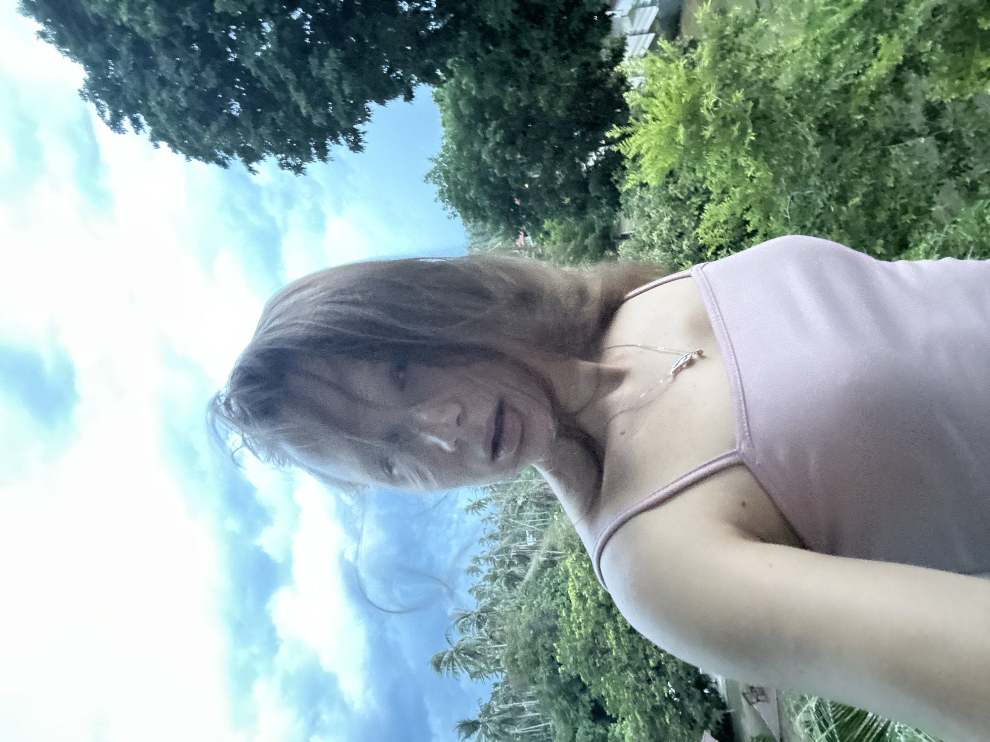 This is a selfie taken outdoors, likely in a tropical location given the presence of palm trees. The subject is a young woman with blonde hair that is somewhat windswept, obscuring parts of her face. She is wearing a light pink tank top with thin straps and a delicate necklace. Her expression appears playful or coy, with her mouth slightly open.
The background is a mix of lush green foliage and a bright blue sky with scattered clouds. The orientation of the image is rotated, suggesting it was taken at an unusual angle, possibly while moving or reaching. The lighting suggests it is daytime, likely midday or afternoon, with bright sunlight illuminating the scene.
The location context provided is Baan Hua Thanon, Thailand, which aligns with the visual cues of tropical vegetation and bright, sunny weather. There is no visible text in the image. The overall mood of the image is casual and relaxed, capturing a moment of personal expression in a natural environment.
