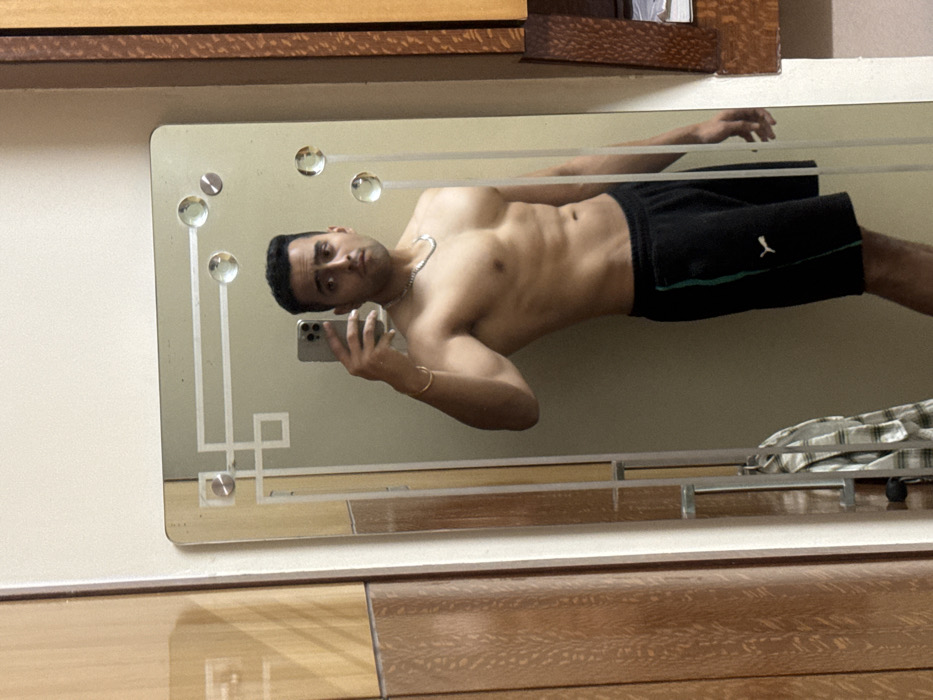 A shirtless man with short dark hair and a silver chain poses in a mirror selfie. He wears black athletic shorts with a green stripe. His physique is toned. The mirror has decorative geometric patterns and round accents. The background features a wooden cabinet or dresser. There is a plaid-patterned cloth on a shelf in the mirror's reflection.