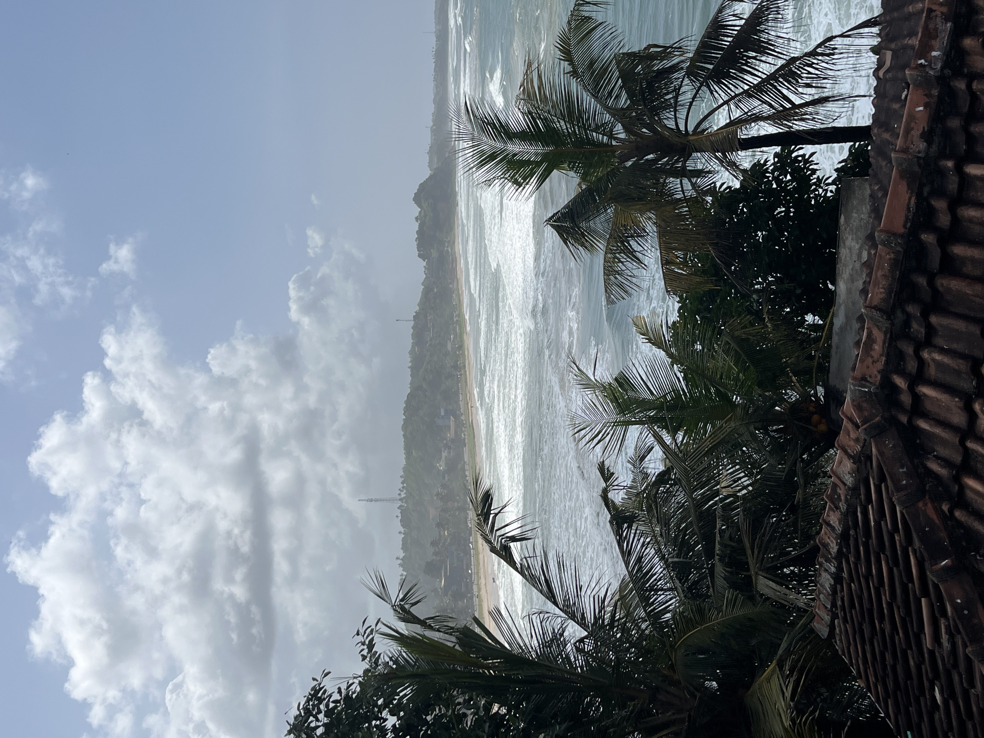 The image captures a coastal scene from a high vantage point, looking out over a beach and the ocean. In the foreground, weathered terracotta roof tiles suggest a building situated above the coastline. Beyond the tiles, lush green palm trees with long, frond-like leaves dominate the middle ground, their dark silhouettes contrasting with the brighter ocean and sky. Some of the palm fronds appear dry and slightly yellowed.
The middle distance features a long, sandy beach that curves along the coastline, dotted with a few buildings and structures that suggest a populated area. A road runs parallel to the beach, and some vehicles are visible. The ocean is active, with numerous waves breaking white and foamy as they approach the shore, indicating a windy or rough sea. The water is a clear blue-green.
In the background, a verdant, rolling hillside rises behind the town. A tall telecommunications tower stands prominently on the crest of the hill. The sky is partly cloudy, with a mix of bright white cumulus clouds and patches of pale blue sky. The overall lighting suggests it is daytime, possibly late morning or early afternoon. The scene is characteristic of coastal Sri Lanka, potentially within or near the city of Matara, known for its beaches and tropical vegetation. No people are clearly discernible in the image.