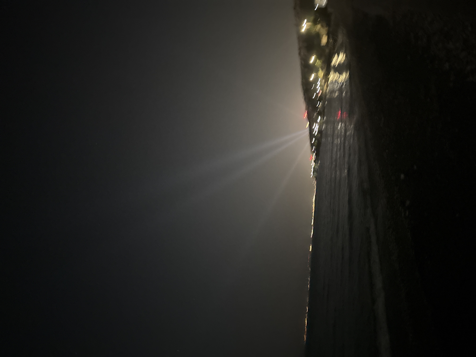 The image depicts a nighttime scene of a coastline, likely within the Agios Tychonas Community of Cyprus. In the foreground, a dark, pebbly beach slopes down to the water. The sea is calm with gentle waves reflecting the distant lights. Along the horizon, a city or town is visible, illuminated by numerous lights that appear blurred due to the low light conditions and possibly motion. Several searchlights or beams of light are visible in the sky, originating from the coastline and pointing upwards into the dark sky. These beams suggest an event or activity taking place on land. The overall atmosphere is dark and nocturnal, with the artificial lights providing the only significant illumination. There are no discernible people or specific objects in the immediate foreground. The environment is consistent with a coastal urban or semi-urban area at night.