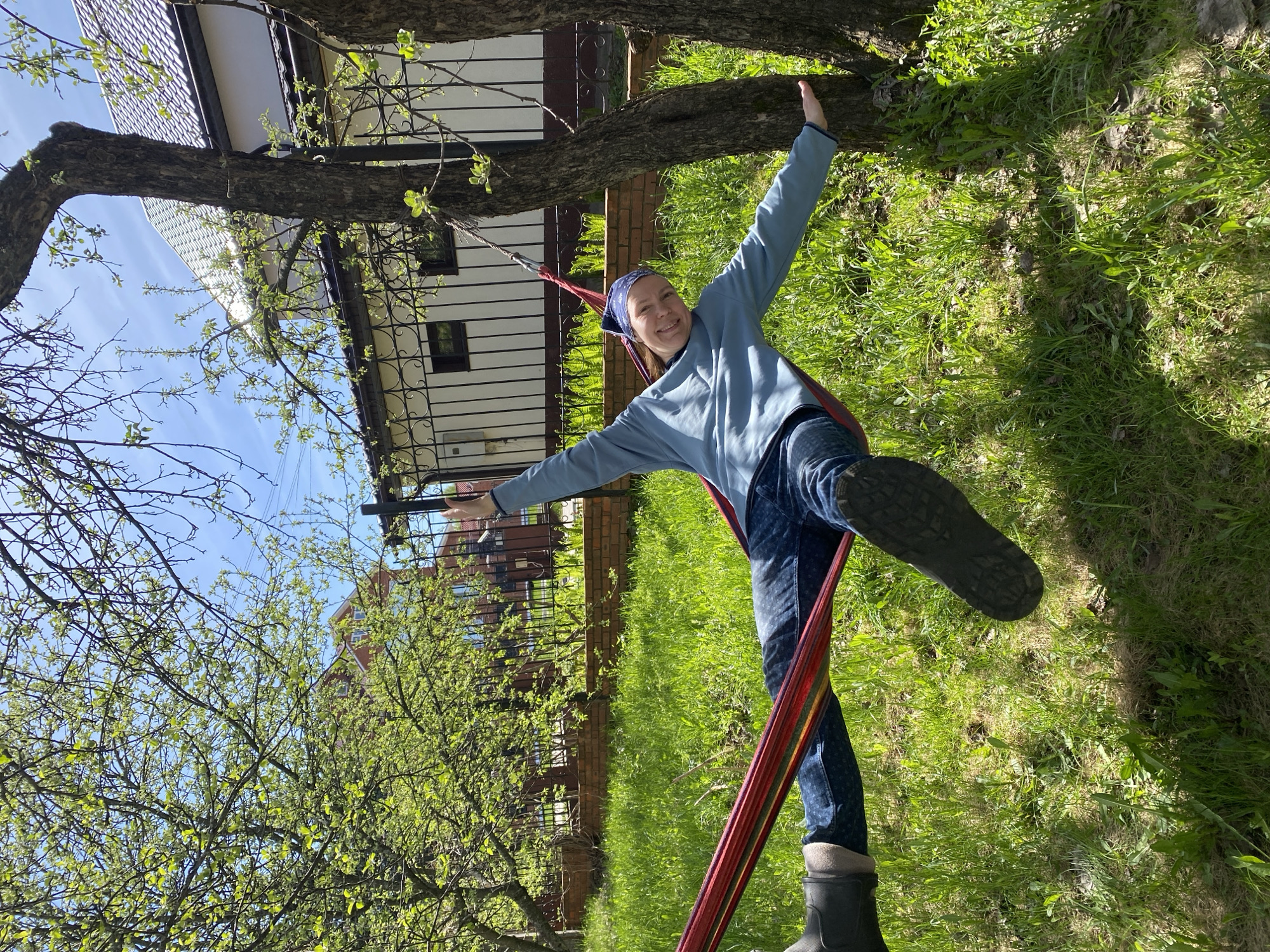 A woman stands in a hammock suspended between two trees in Прикамский, Russia. She wears a blue hooded shirt, blue jeans, a blue headscarf, and black rubber boots. Her arms are outstretched, touching the tree trunks. The background includes green grass, trees with budding leaves, a brick wall, a fence, and a white house with dark windows. The sky is visible through the trees.