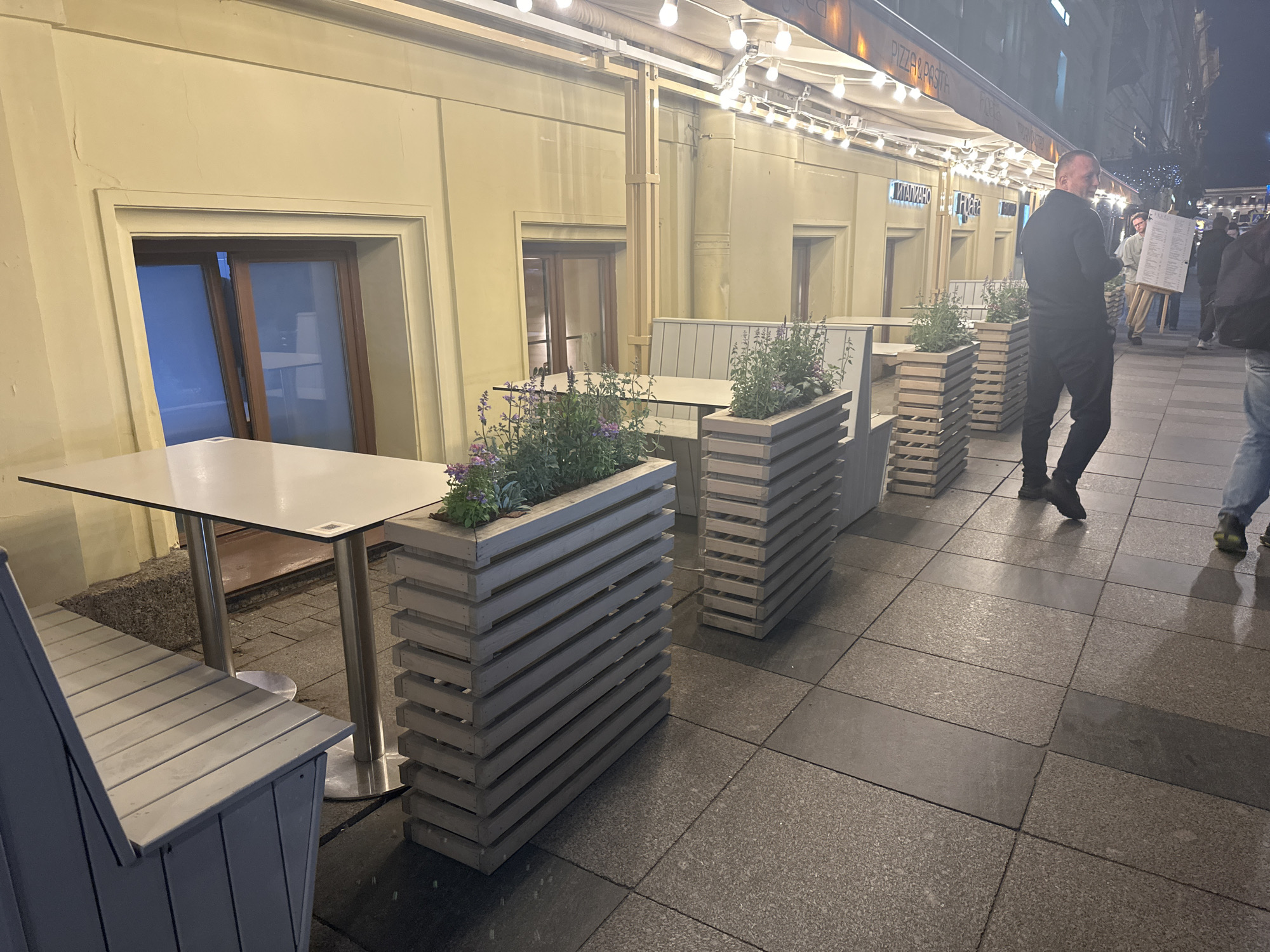The image depicts an outdoor dining area along a street in Saint Petersburg, Russia, likely in the evening. The scene is illuminated by overhead string lights, suggesting a "Pizza & Pasta" restaurant or similar eatery. Tables and benches are arranged outside the building. Each table has a planter with flowers on one side. Several people are walking on the sidewalk nearby, including a man in a black suit. The restaurant's signage is visible in the background, although somewhat blurred. The ground is paved with dark gray tiles. There are doors and windows on the side of the building.