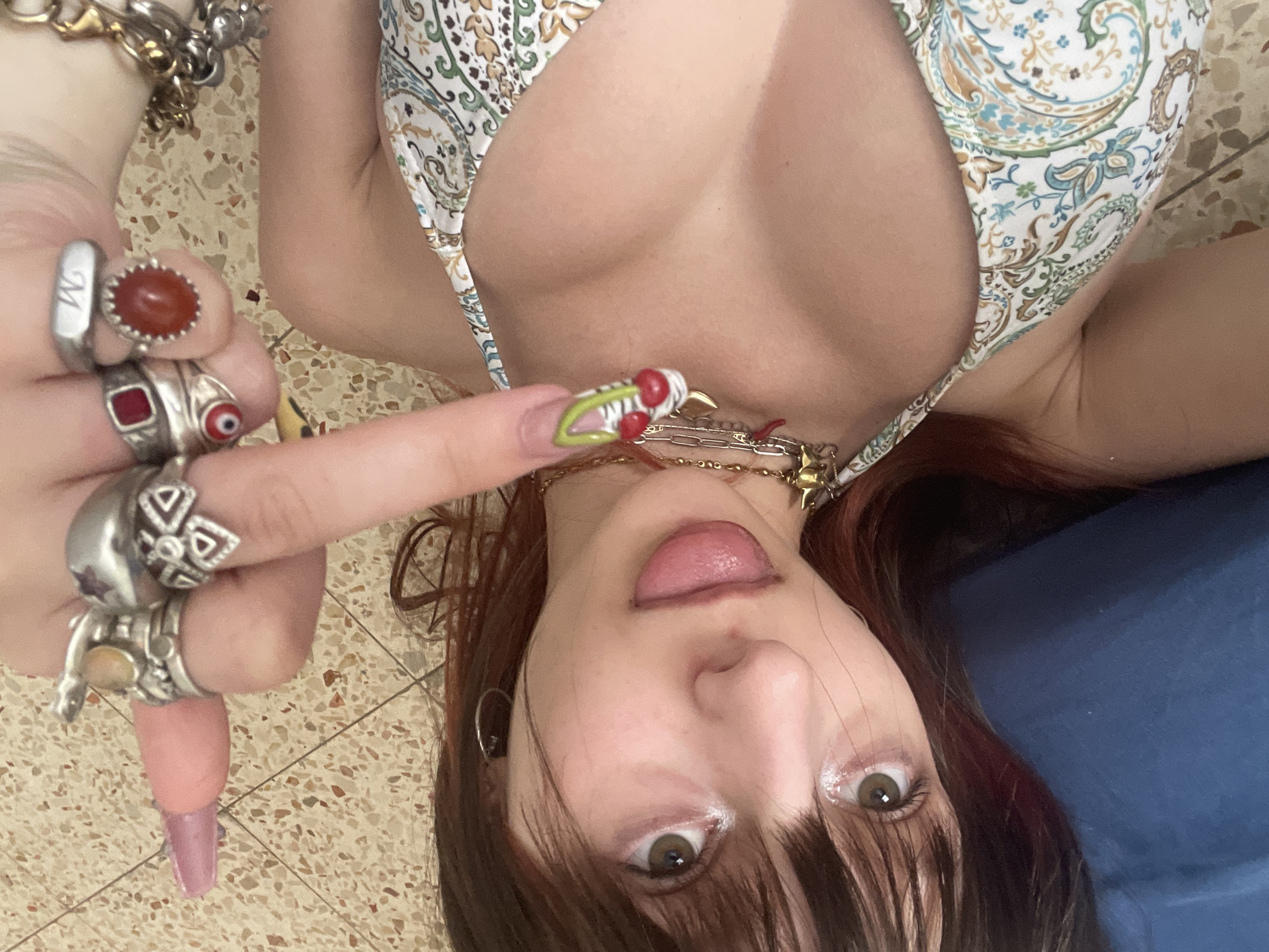 A woman is looking upwards towards the camera. Her tongue is slightly sticking out. She is wearing a patterned bikini top. Her left hand is prominently displayed, adorned with multiple rings. She is wearing a number of necklaces. The background appears to be a tiled surface. The other background item is a blue surface. There is no evidence to indicate the city of Petah Tikva, Israel.