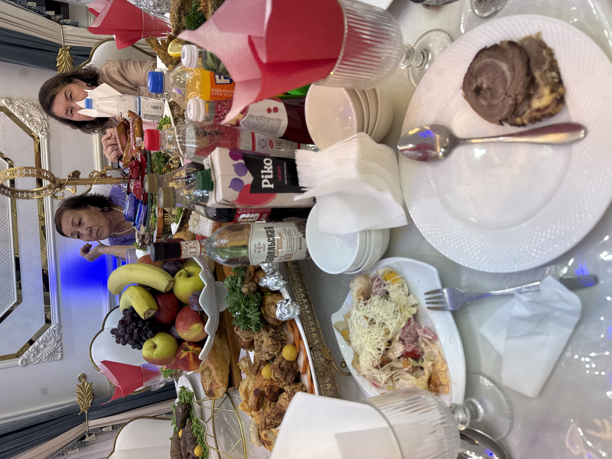 The image depicts a table laden with food and drinks, likely a meal or celebration. Two women are seated at the table, one in the foreground and another further back. The woman in the foreground is wearing a tan-colored top and looking towards the left of the frame. The woman in the background, to the left, is wearing a purple top and also looking to the left. The table is set with plates, cutlery, glasses, and various food items, including a fruit bowl with bananas, grapes, apples, and other fruits, a platter of fried meats and pastries, a salad, and several bottles of drinks, including what appears to be a bottle of vodka. Red napkins are placed in decorative holders. The setting suggests an indoor environment, possibly a home or restaurant, with ornate wall decorations and lighting visible in the background. The presence of a wide variety of food and drinks indicates a festive occasion. The image appears to be taken in Şardara, Kazakhstan, given the provided context.