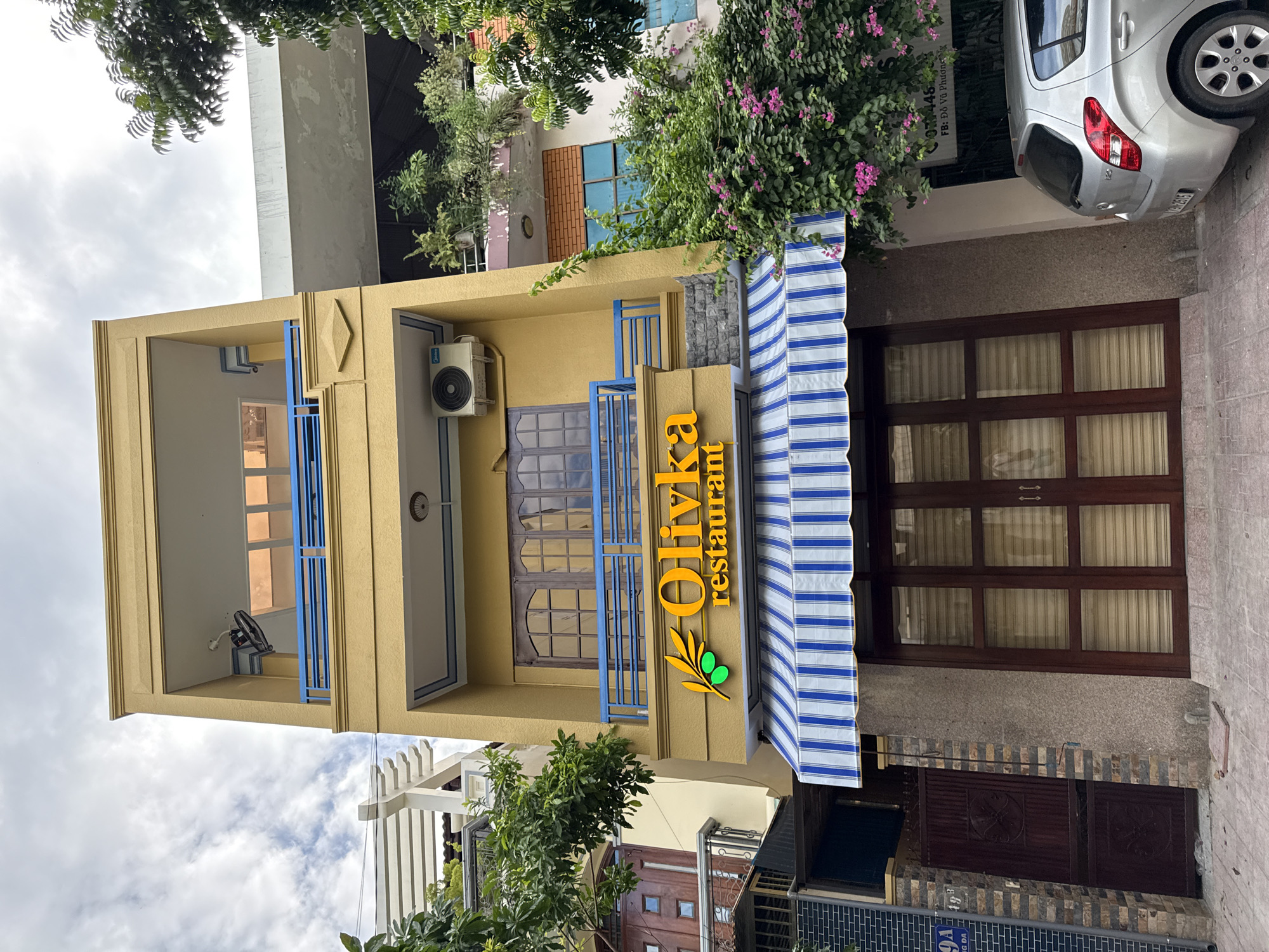The image shows a three-story yellow building with a blue railing on the balconies. On the second floor, there is a sign with the text "Olivka restaurant" in orange lettering, accompanied by a green olive branch logo. Below the sign is a blue and white striped awning, and beneath that, large wooden double doors. To the left of the restaurant entrance, there is a dark red metal gate with decorative patterns. To the right of the building, a silver Hyundai car is parked on the street, partially visible. Green trees and pink bougainvillea flowers are present on the right side of the building, and a white air conditioning unit is visible on the second floor. The sky is cloudy. The street number "49A" is visible on a sign to the left of the gate. The building appears to be located on a street in Nha Trang, Vietnam, likely in a commercial or residential area.