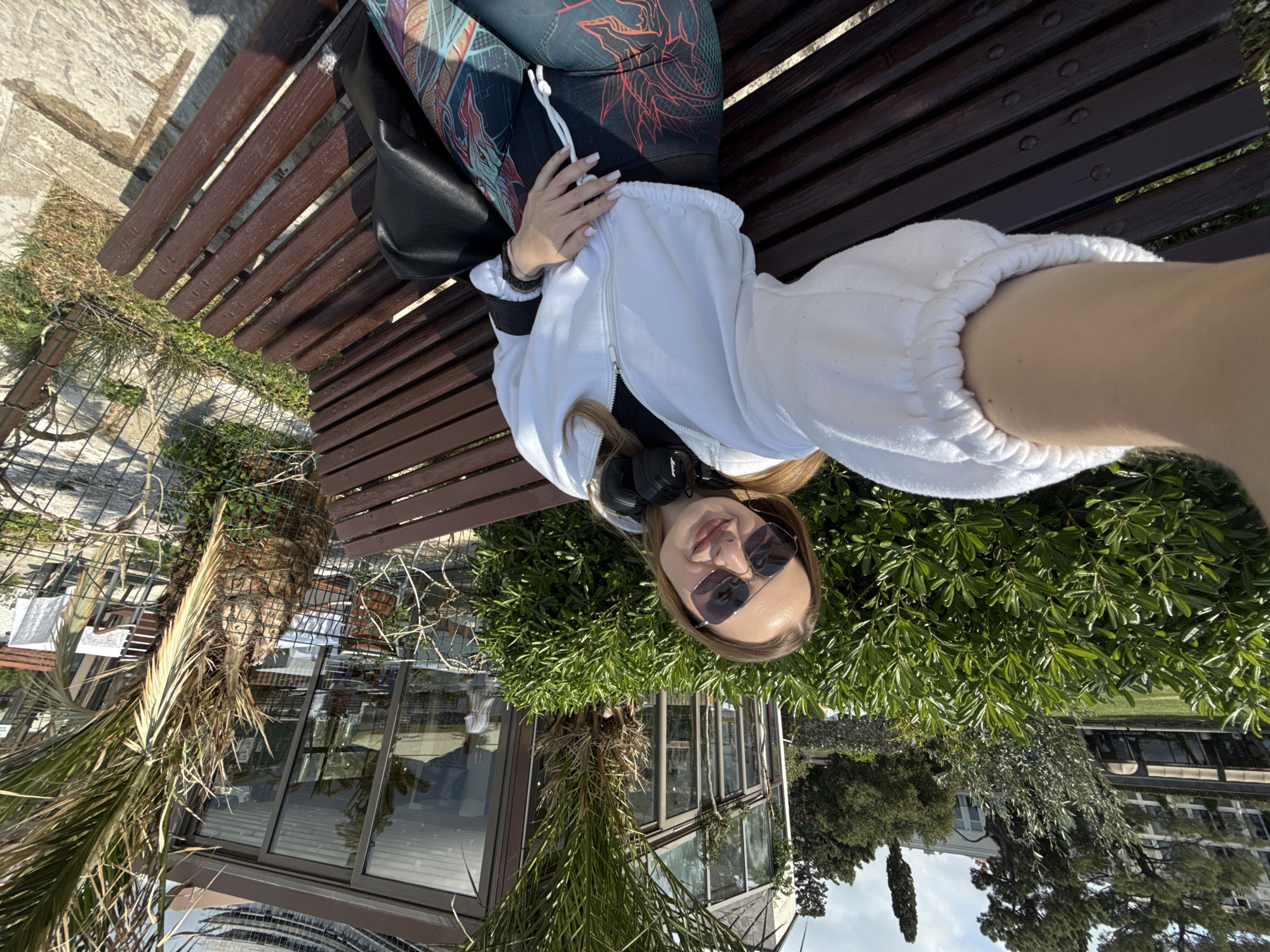 The image depicts a young woman taking a selfie while lying comfortably on a wooden slatted bench under bright daylight in Budva, Montenegro. She is positioned with her head towards the top of the frame, looking directly into the camera with sunglasses partially obscuring her eyes and a relaxed expression. Her light brown hair frames her face, and a pair of black Marshall headphones rests around her neck.
She is dressed in casual attire, including a white zippered hoodie with ribbed cuffs and a dark top underneath. Her leggings feature a distinct pattern of red, orange, and blue intricate designs. A black bag is visible near her hip, and her hand, with light-colored manicured nails, rests gently on her stomach.
The setting is outdoors, characterized by abundant green vegetation. A prominent palm tree is visible to the left of the bench, and various other green bushes and trees fill the mid-ground and background. Behind the foliage, modern glass-fronted buildings, possibly reflecting the sunny sky, are discernible. The ground around the bench consists of light-colored paving and some patches of grass. The clear sky and strong sunlight indicate a warm, pleasant day.