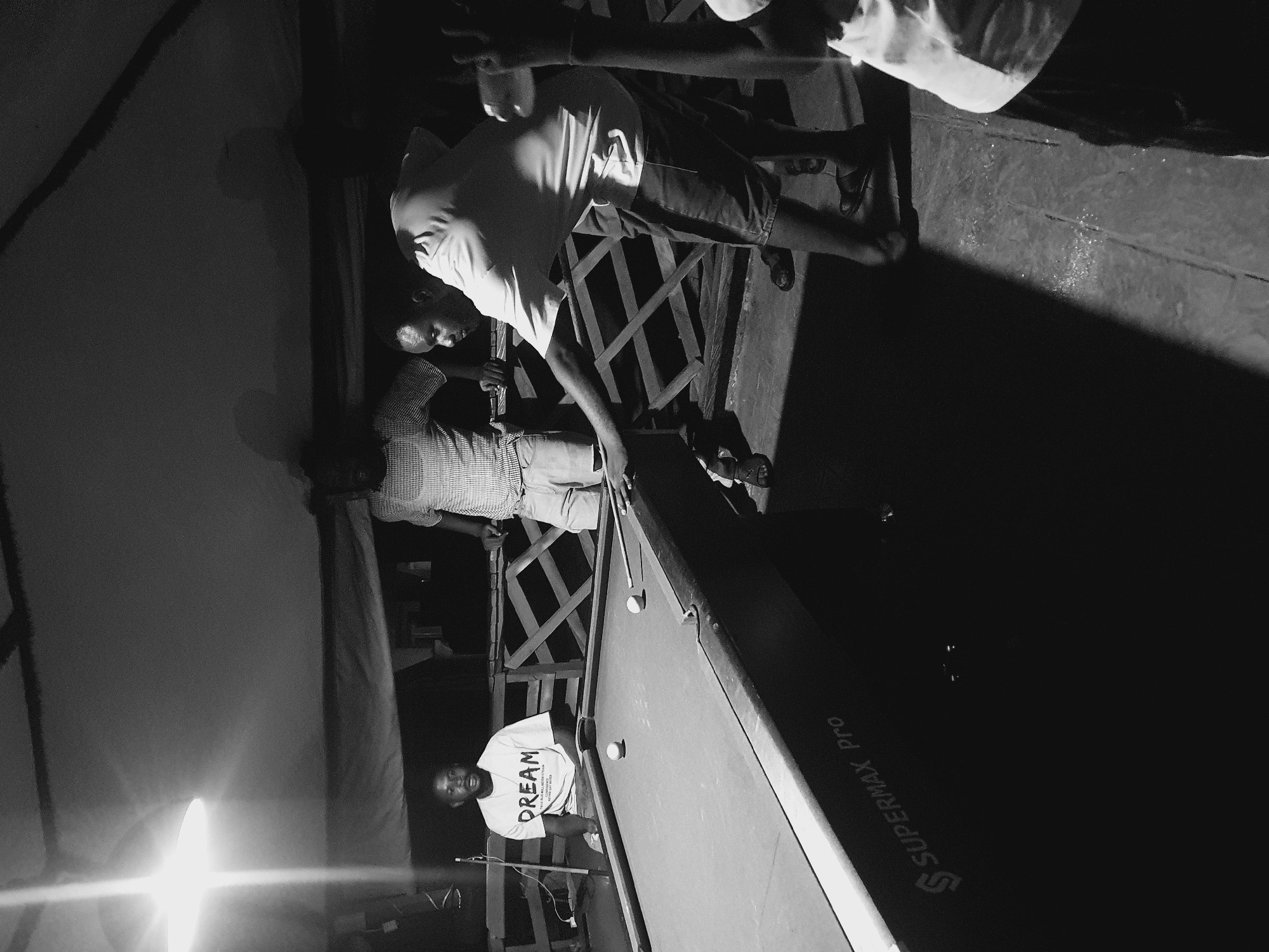 The image depicts a group of individuals engaged in a game of pool at night, under artificial lighting, likely at an outdoor establishment in Lokoja, Nigeria. A pool table, labeled "SUPERMAX Pro," dominates the foreground. Two white billiard balls are visible on the green felt. One man, wearing a t-shirt with "DREAM" printed on it, is seated and observing the game. Another man, in a checkered shirt, stands behind him, also watching. A third man, leaning over the pool table, is actively playing, with a cue stick in his hand. His posture suggests he is about to strike a ball. The background reveals wooden railings and structures, indicating an open-air setting. The overall lighting is dim, with a bright overhead light casting strong shadows. The scene is captured in black and white, emphasizing the textures and forms within the frame.