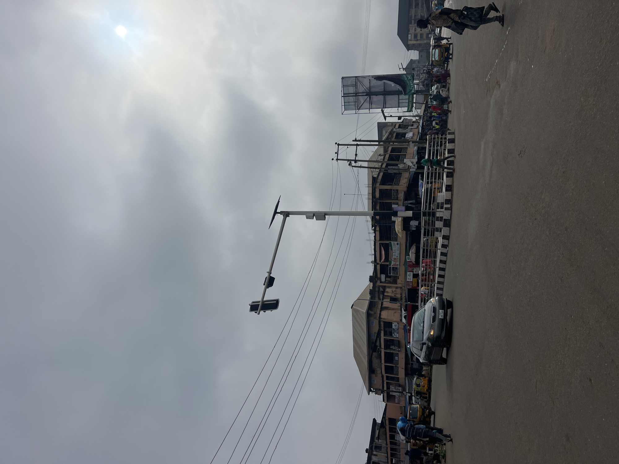 The image depicts a street scene in Nkpor, Nigeria, under an overcast sky with the sun faintly visible through the clouds. A four-way intersection is the central focus, with a grey sedan driving through it. Traffic lights hang over the intersection, indicating a controlled traffic flow. Buildings line the sides of the street, featuring multiple stories with balconies and shops on the ground level. Several people are visible, some walking, others standing, and one person is seen crossing the street in the foreground. Various vehicles, including at least two yellow tricycles, are present in the background. Electrical wires crisscross above the street, supported by poles. A large billboard stands on the right side of the image. The overall environment suggests a busy urban commercial area.