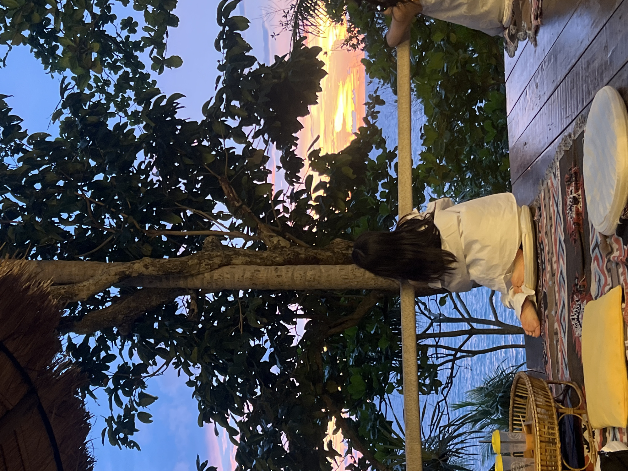 A person sits on a wooden deck overlooking the ocean at sunset in Karon, Thailand. The person is facing away from the camera, wearing a white shirt and pants, and is sitting on a cushion. The deck is furnished with a woven tray holding two drinks, a rug, and pillows. Trees and foliage frame the view of the ocean and the colorful sunset. The sky is a blend of blue, pink, and orange hues. Another person's arm is visible on the right side of the image. The overall scene depicts a serene moment in a tropical setting.
