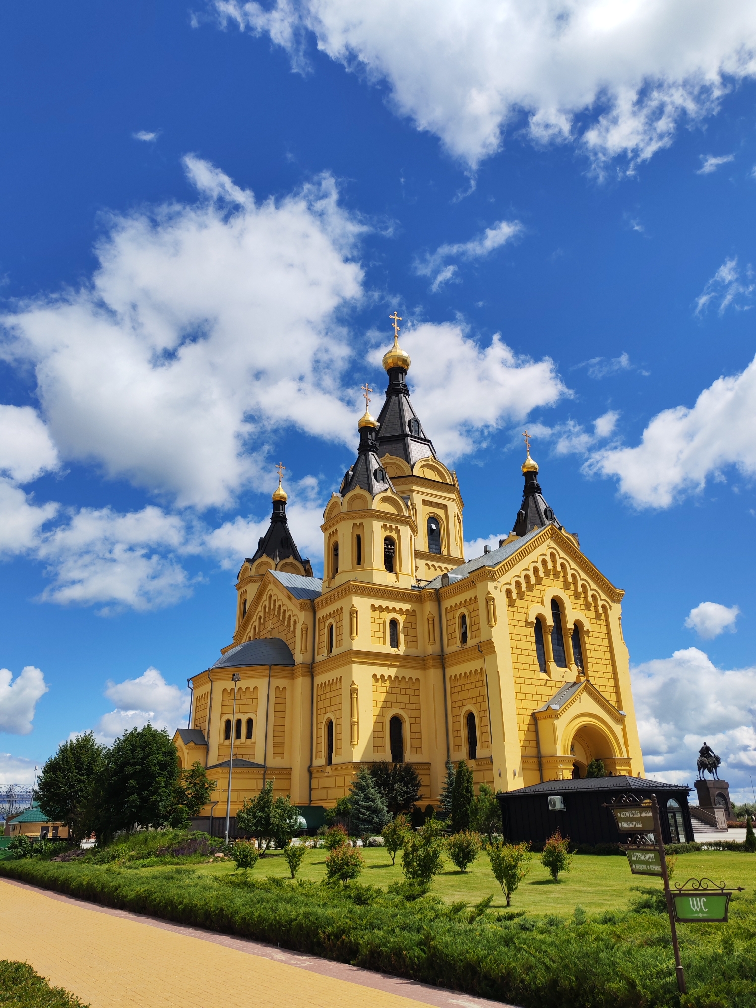 The image displays a bright yellow Orthodox cathedral with multiple black domes topped with gold crosses, set against a vivid blue sky with scattered white clouds. The cathedral features intricate architectural details, including arched windows and decorative cornices. In the foreground, a meticulously manicured green lawn is bordered by trimmed hedges. To the left, a pathway paved with light brown interlocking stones leads the eye towards the cathedral. A dark, rectangular building with a black roof is situated near the cathedral entrance, adorned with several signs. To the right of the cathedral, a distant equestrian statue is visible. The scene is bathed in natural sunlight, suggesting an outdoor daytime setting. This is the Alexander Nevsky Cathedral, located in Nizhny Novgorod, Russia.